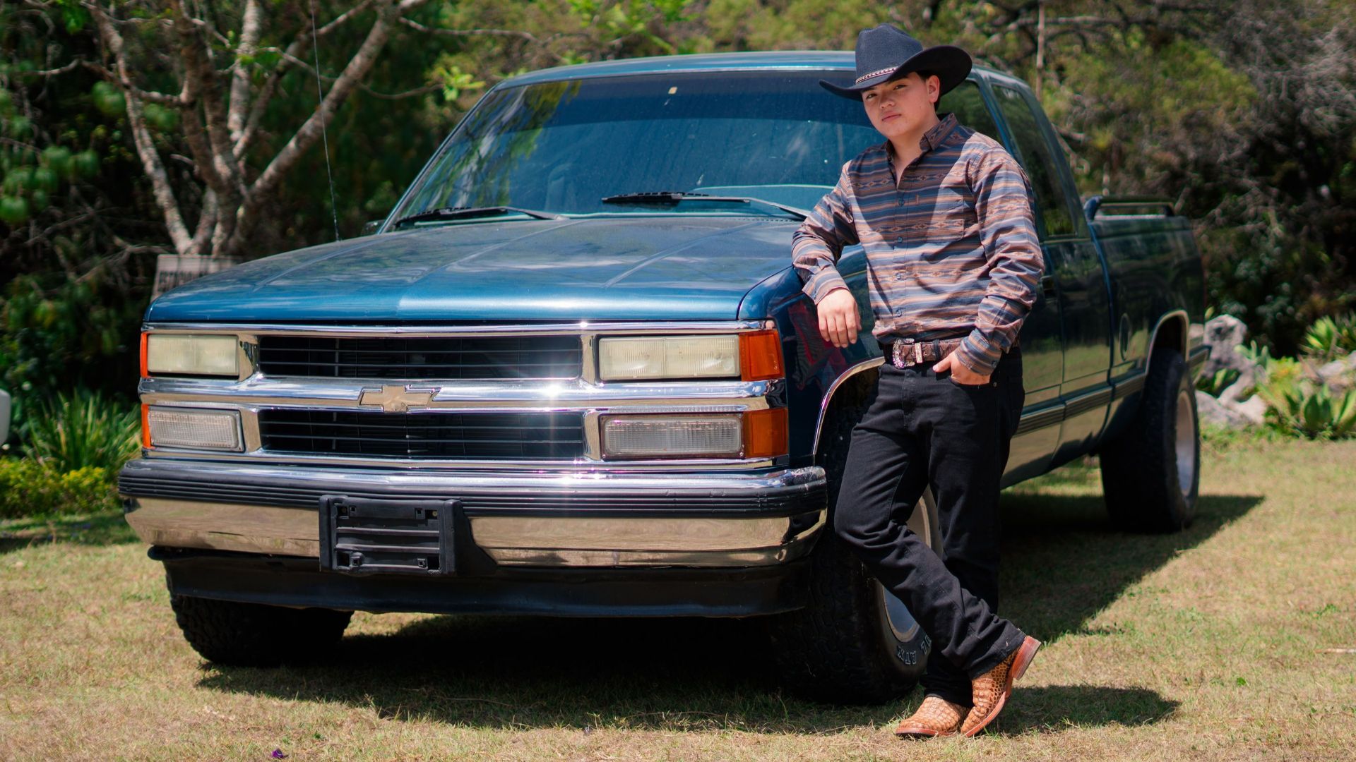 A cowboy in a hat relaxes against a classic blue truck outdoors in a sunny field.