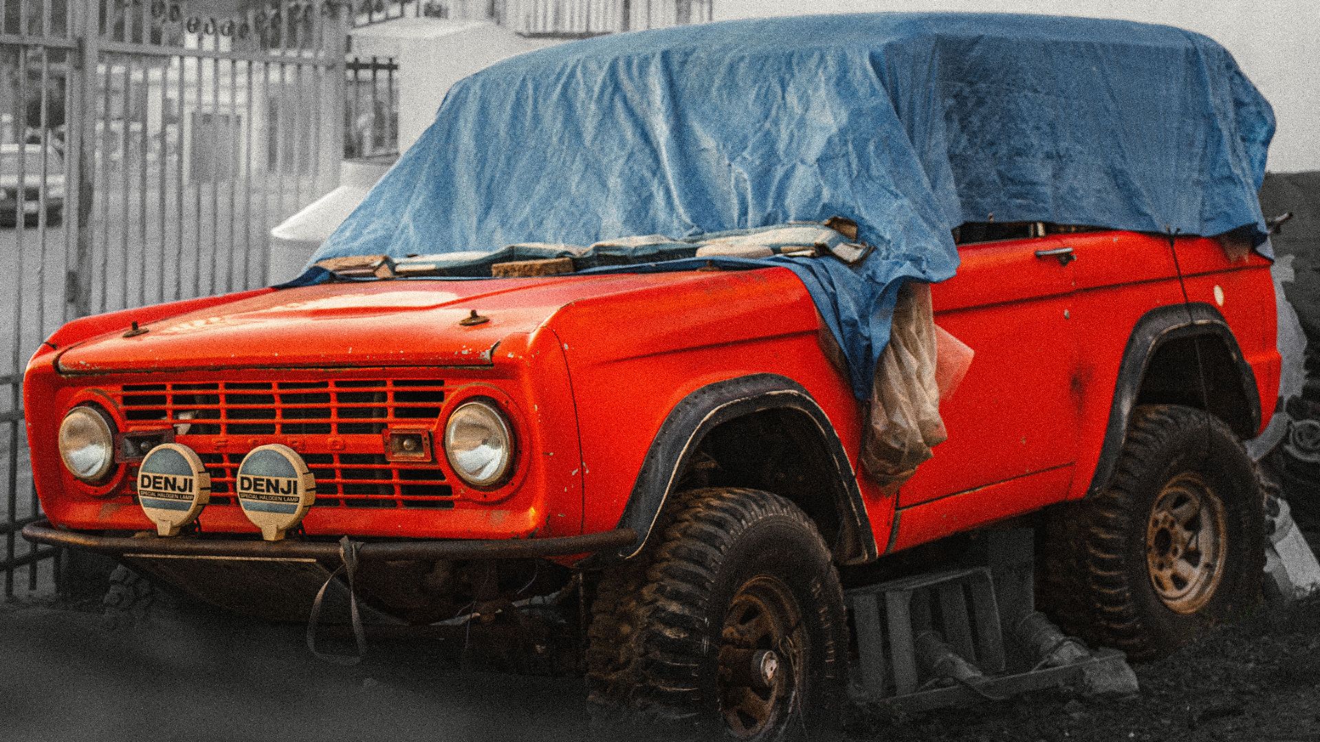 A classic red SUV covered with a blue tarp, parked outdoors, showcasing vintage charm.
