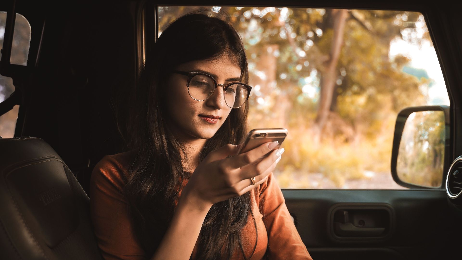 A young woman wearing glasses and casual clothing relaxes in a car, using her phone on a sunny day.