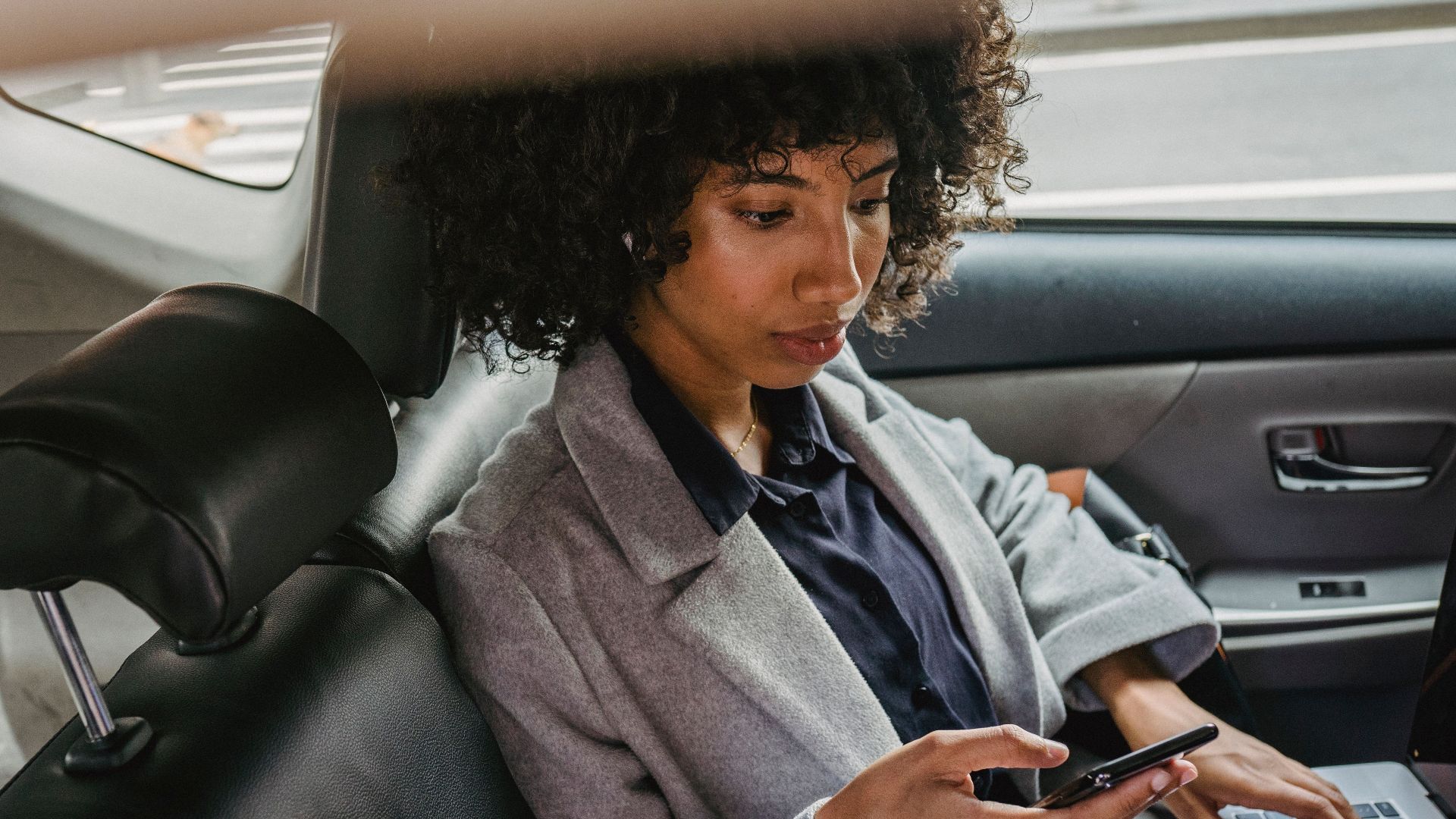 Woman multitasking with phone and laptop in car backseat, balancing work and travel.
