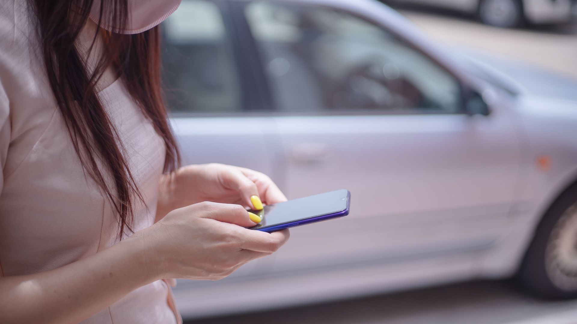 A close-up of a woman texting on a smartphone near a parked car outdoors.