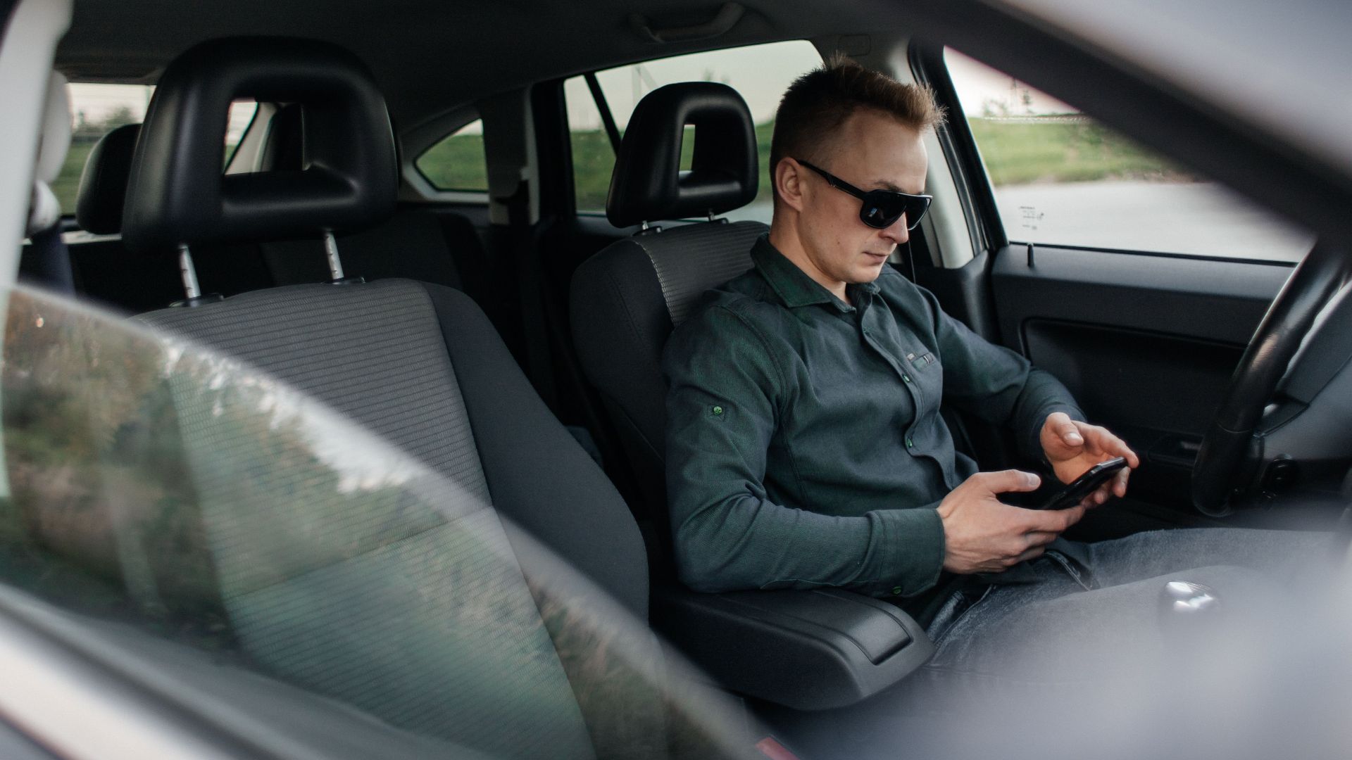 Young man using smartphone inside a parked car, wearing sunglasses.