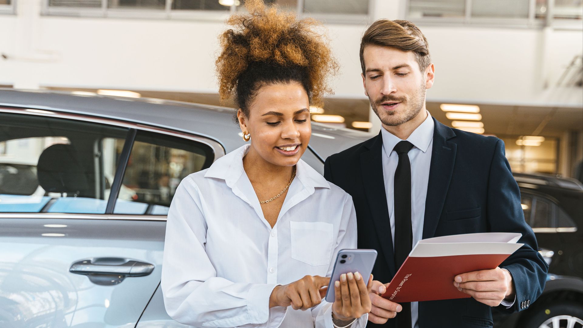 Two professionals discussing business while looking at a mobile phone in a car showroom.