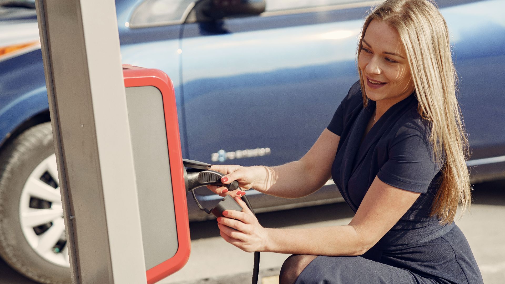 Side view of positive female in casual dress sitting near station on street charging electric vehicle for continuation trip