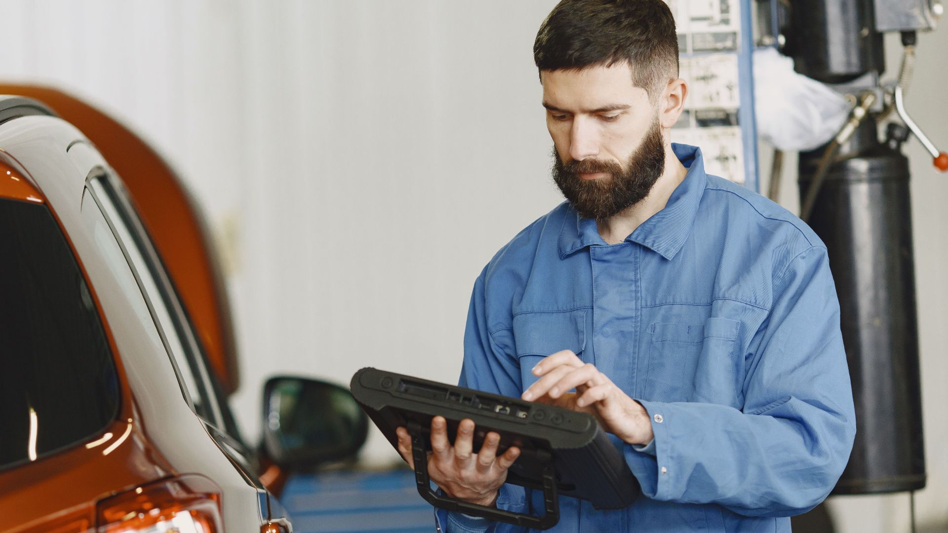 A mechanic in coveralls using a diagnostic tool at an auto repair shop beside a car.