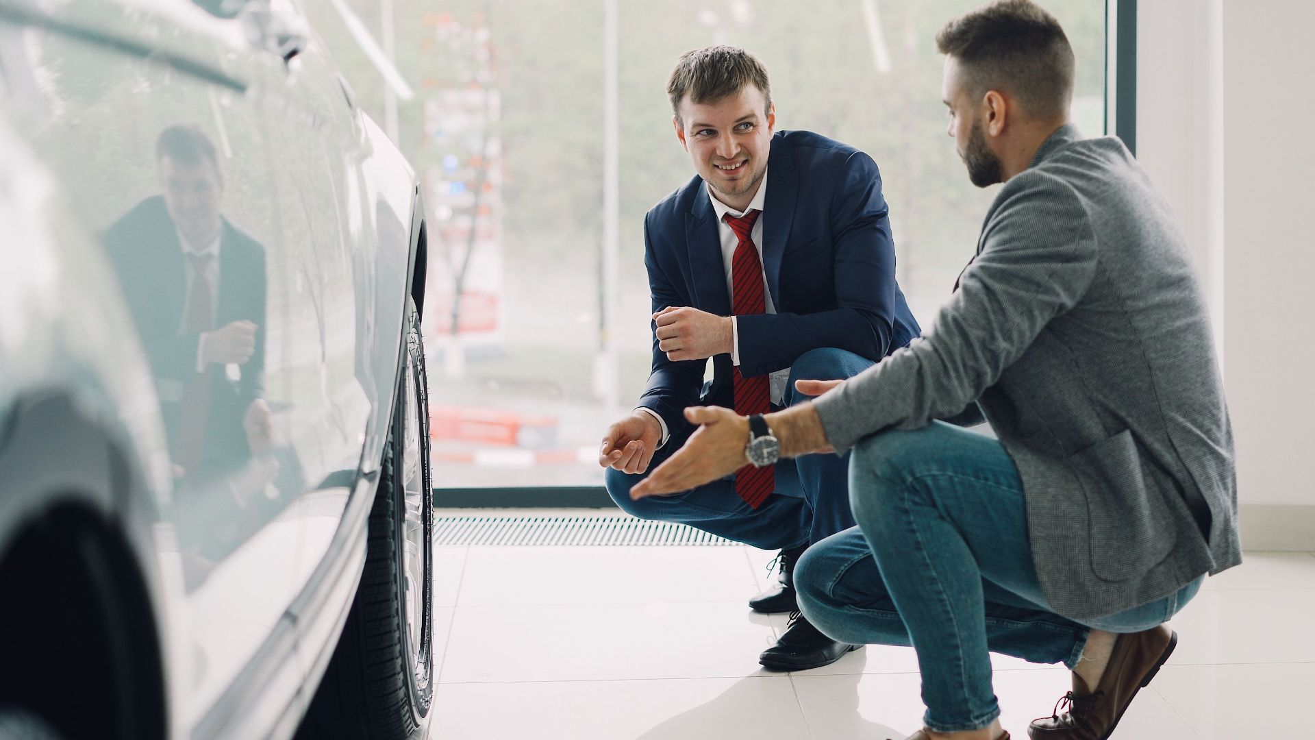 Two men discussing car features in a showroom, kneeling near a vehicle.