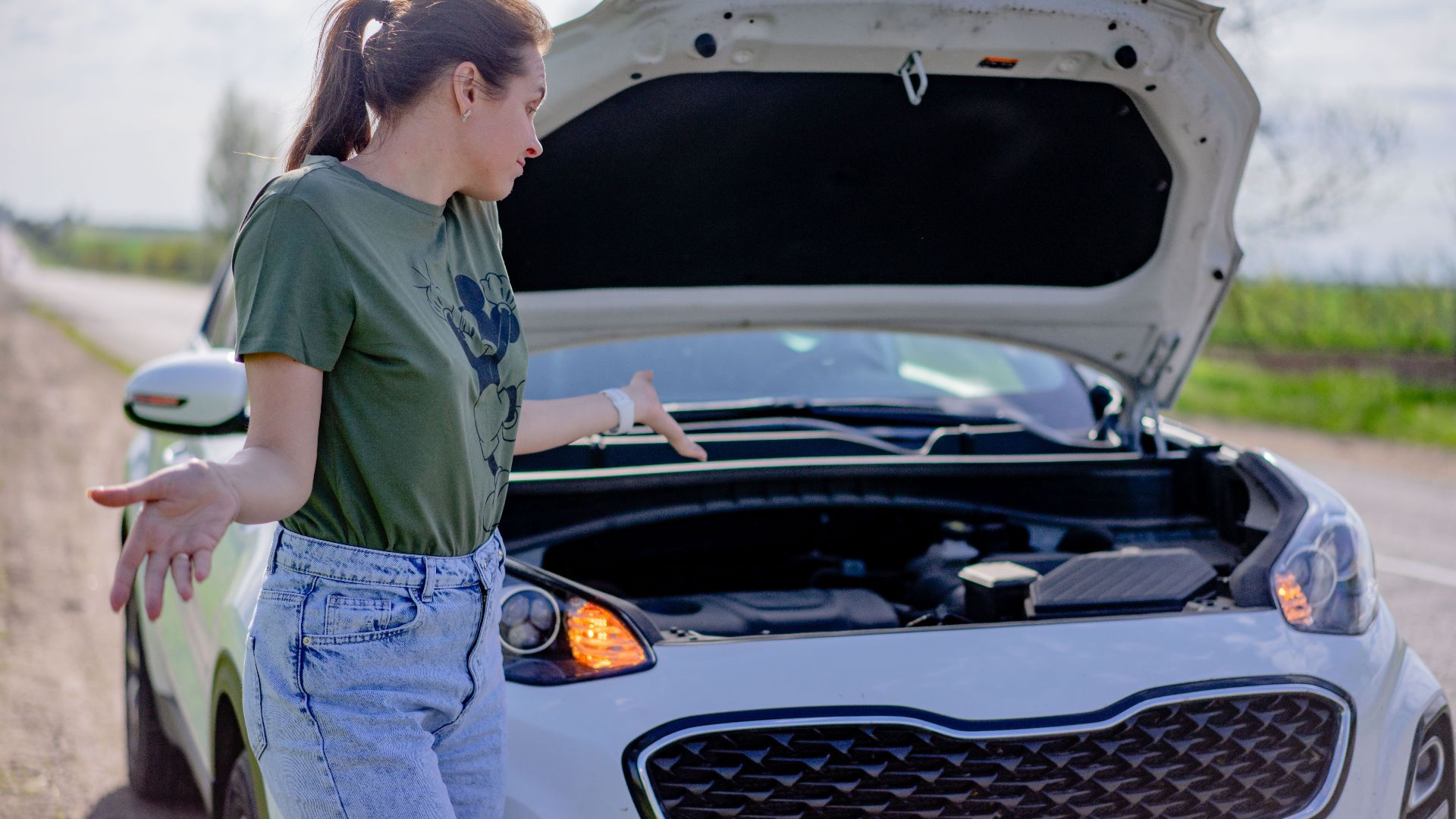 A woman looks puzzled by her broken car with an open hood on a rural road.