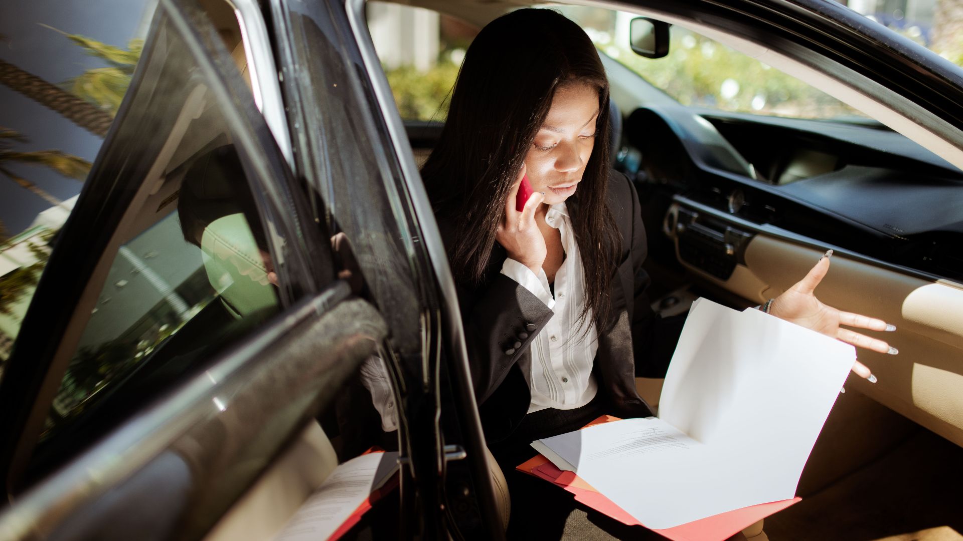 A businesswoman multitasking with phone and documents while seated in a car.