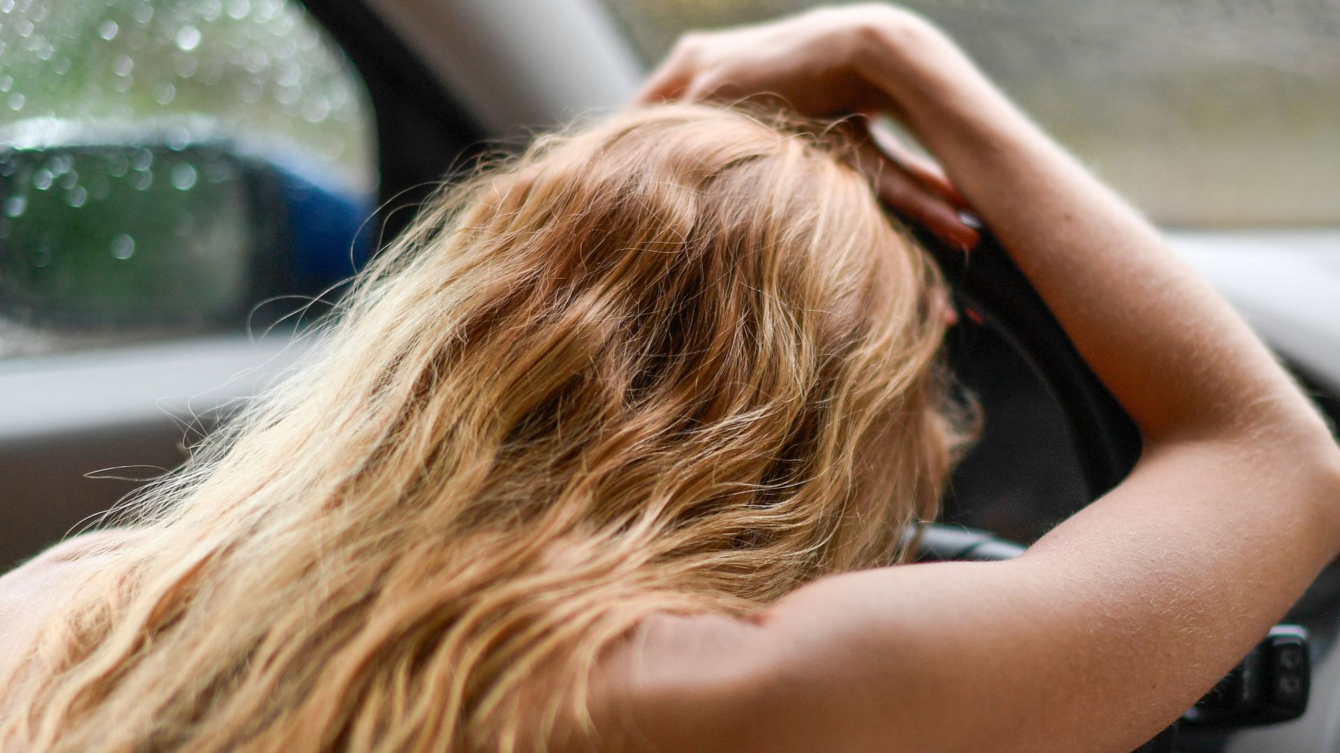 A woman with blonde hair leans on a steering wheel inside a car, creating a contemplative and serene mood.