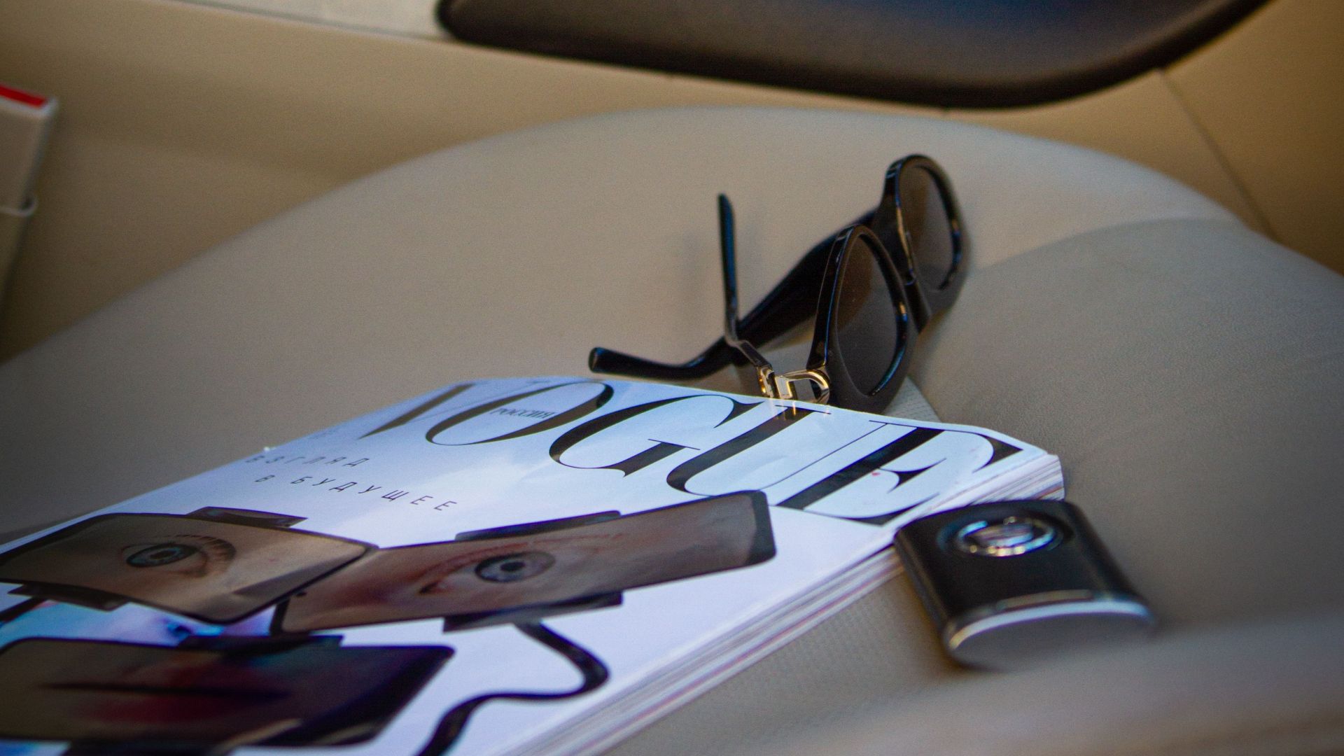Close-up of a magazine and sunglasses on a car seat with a person using a smartphone in the background.