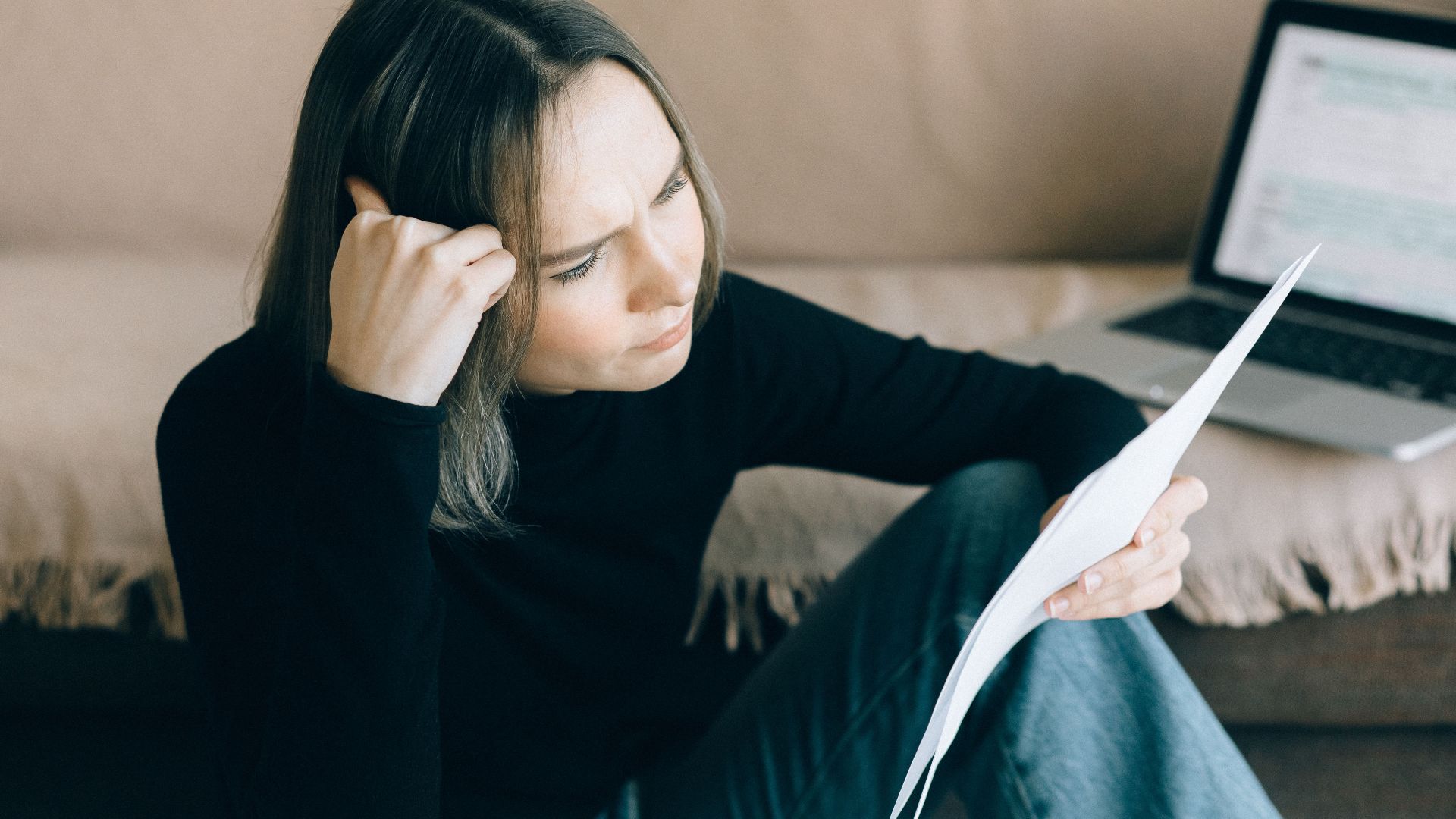 Focused woman analyzing papers with a laptop open, symbolizing thoughtful consideration.