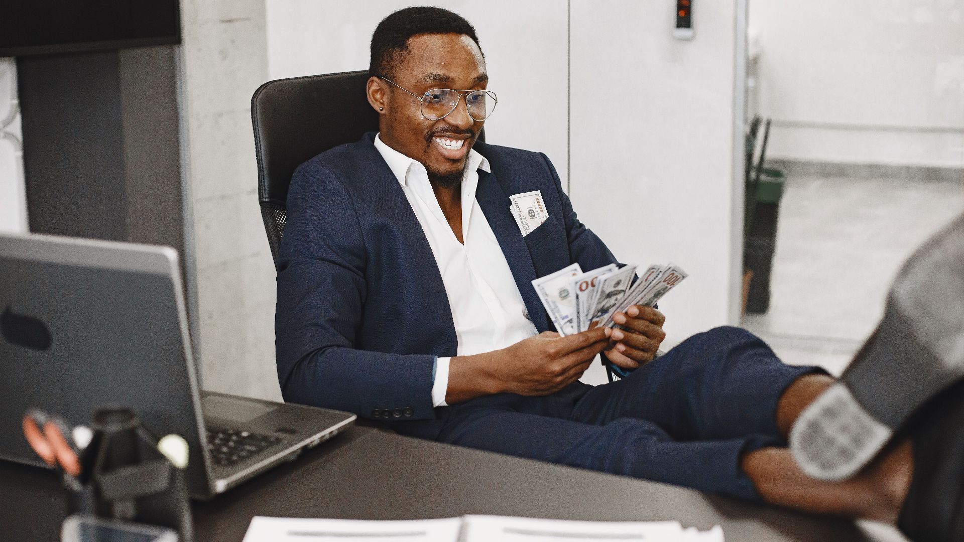 Businessman in a suit counts cash with a smile, relaxing in a modern office setting.