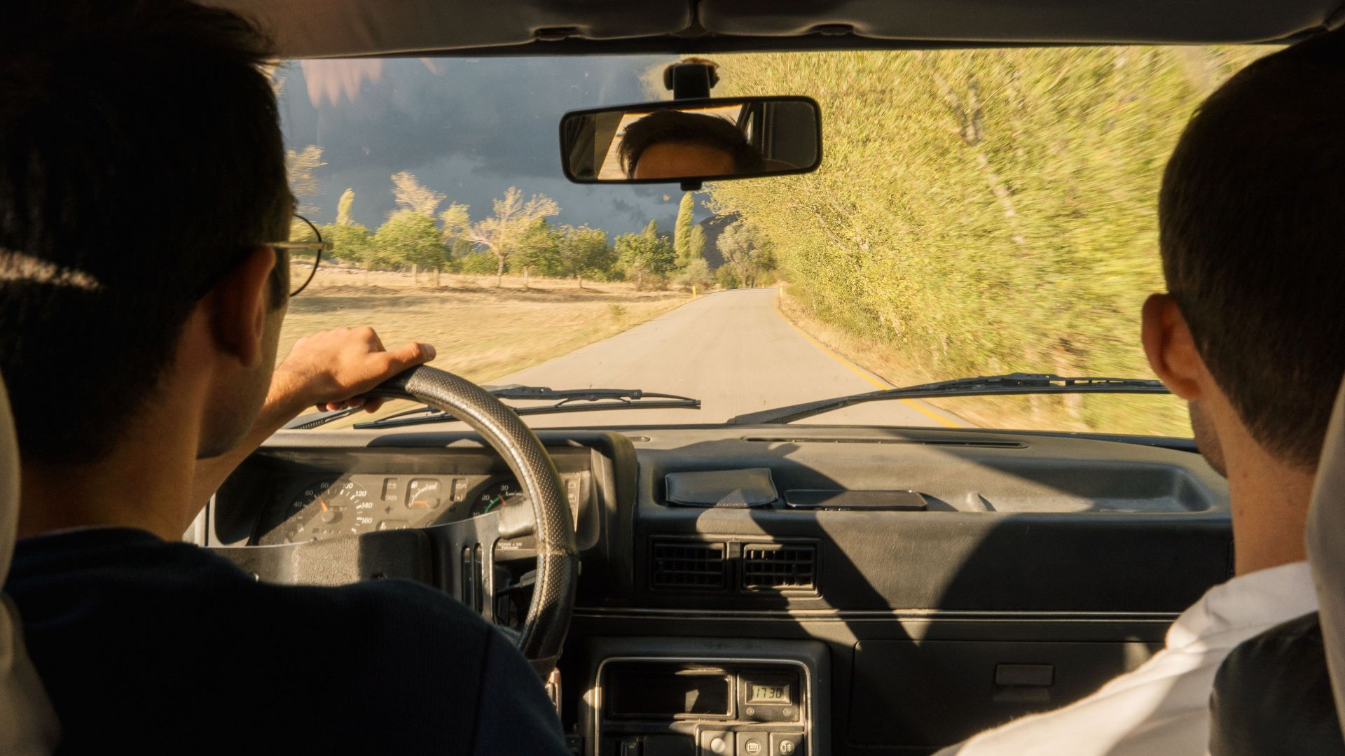 Two adults driving through scenic countryside near Merzifon, Türkiye, in a vintage car.