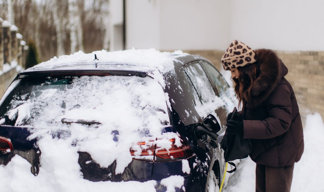 Woman connecting charging cable to modern electric vehicle covered in snow during cold winter weather, promoting green energy