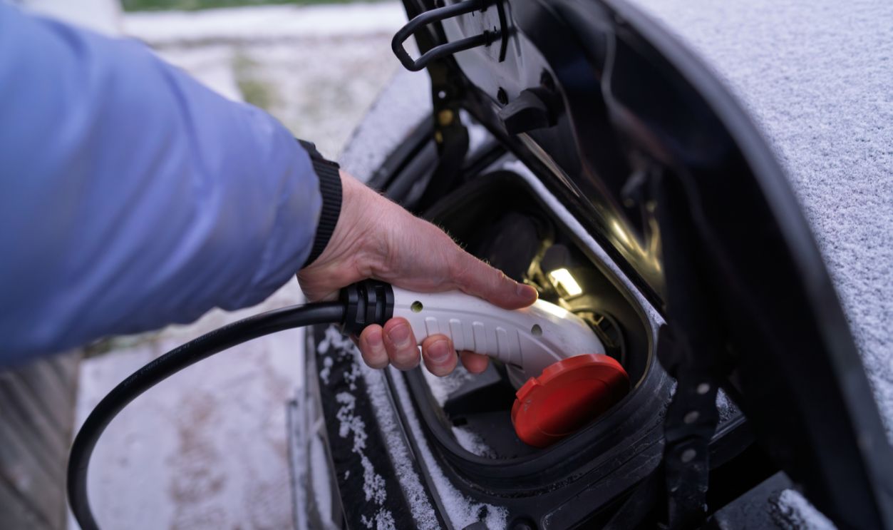 Hand connecting electric car charger to vehicle charging port in winter weather, white EV plug with red cap on snow-covered automobile, green technology and sustainable mobility concept