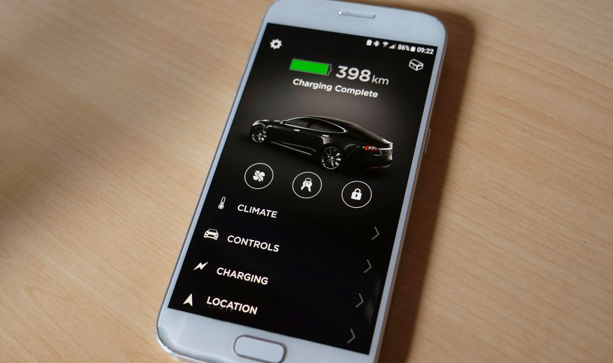 TESLA AUTONOMOUS CAR, March 2018 - CLOSE UP: White smartphone lying on the wooden desk signalling the Tesla car is fully charged. Cool cell phone application controlling the futuristic car's battery.