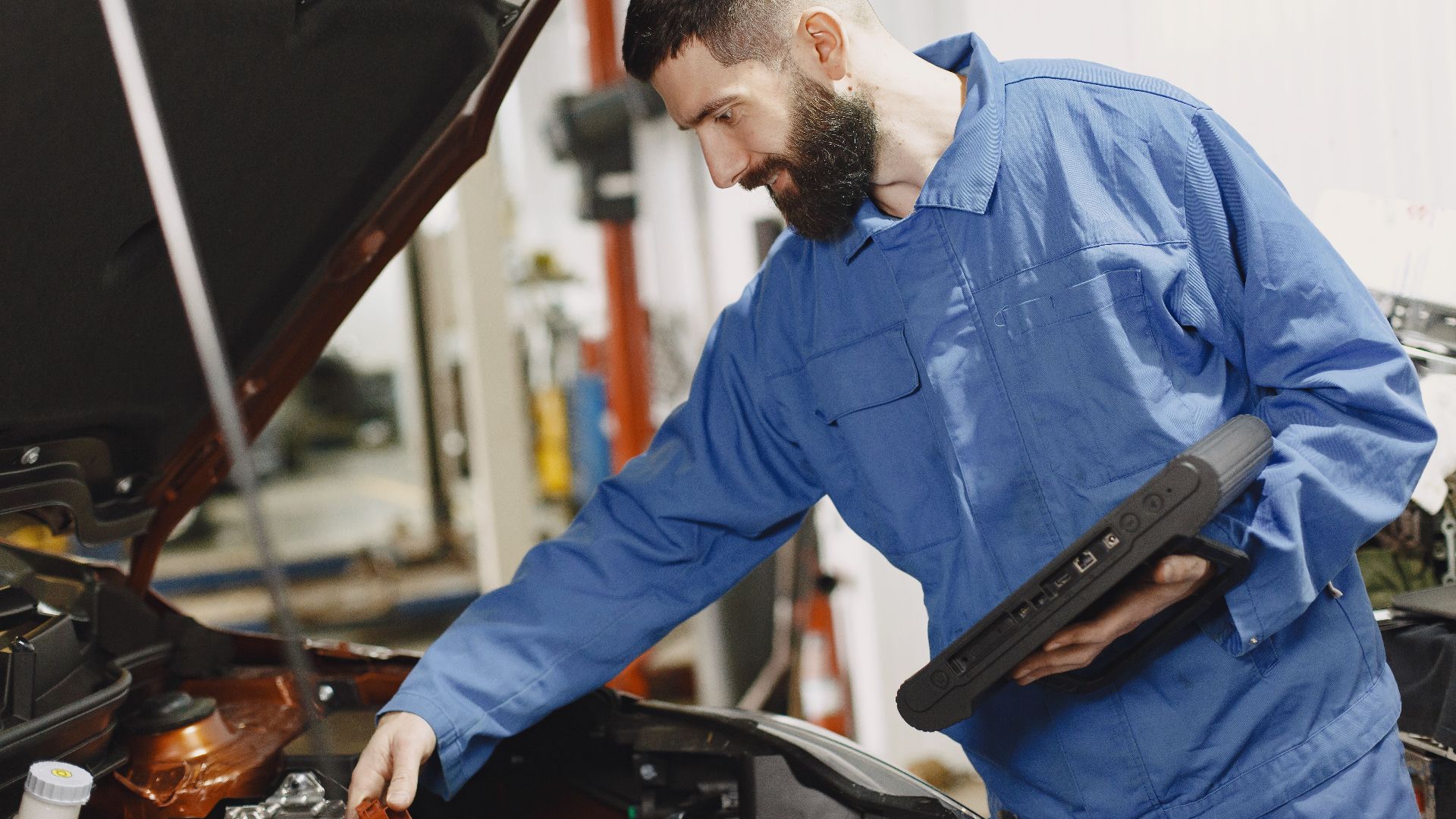 An auto mechanic in blue coveralls inspecting a car's engine in a garage.