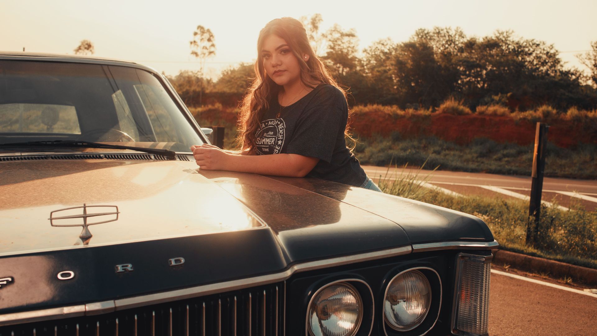 A young woman poses by a classic Ford car during sunset on a rural road.