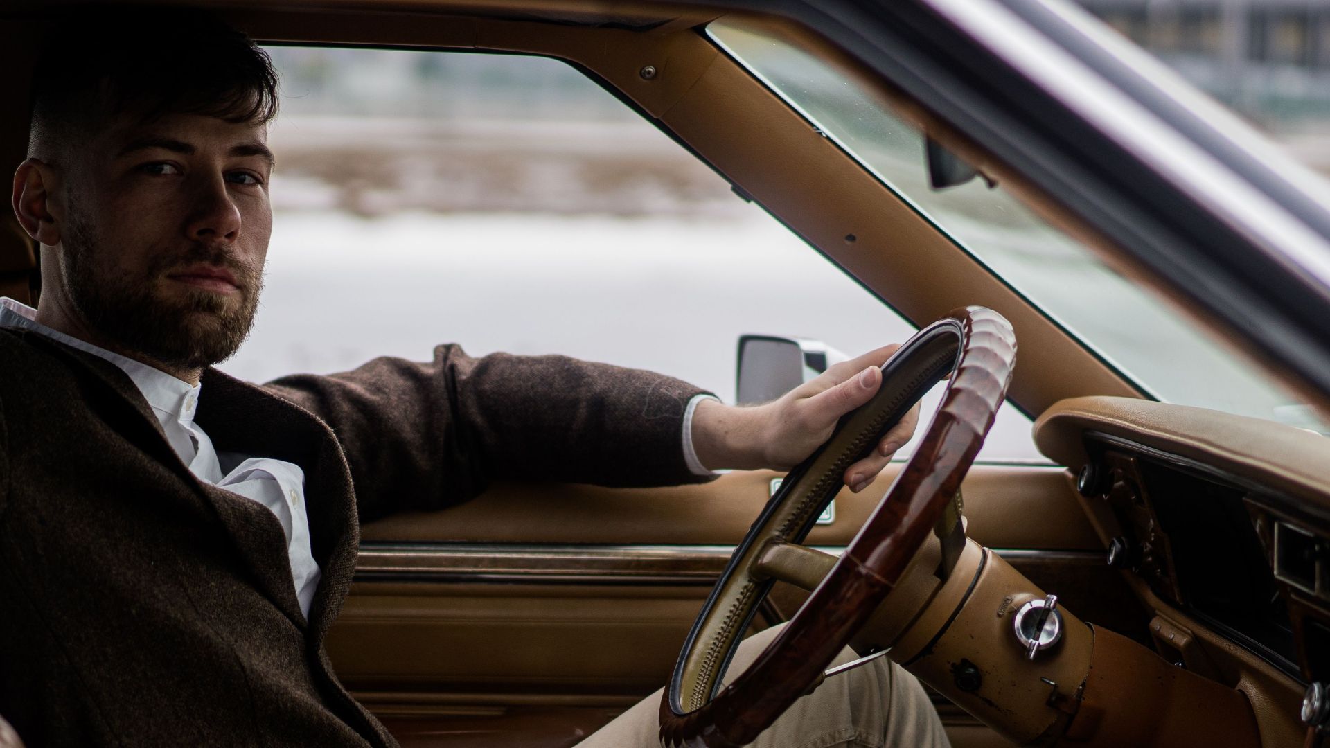 Side view of confident adult male driver in elegant outfit sitting in vehicle with hand on steering wheel while looking at camera on street in daytime