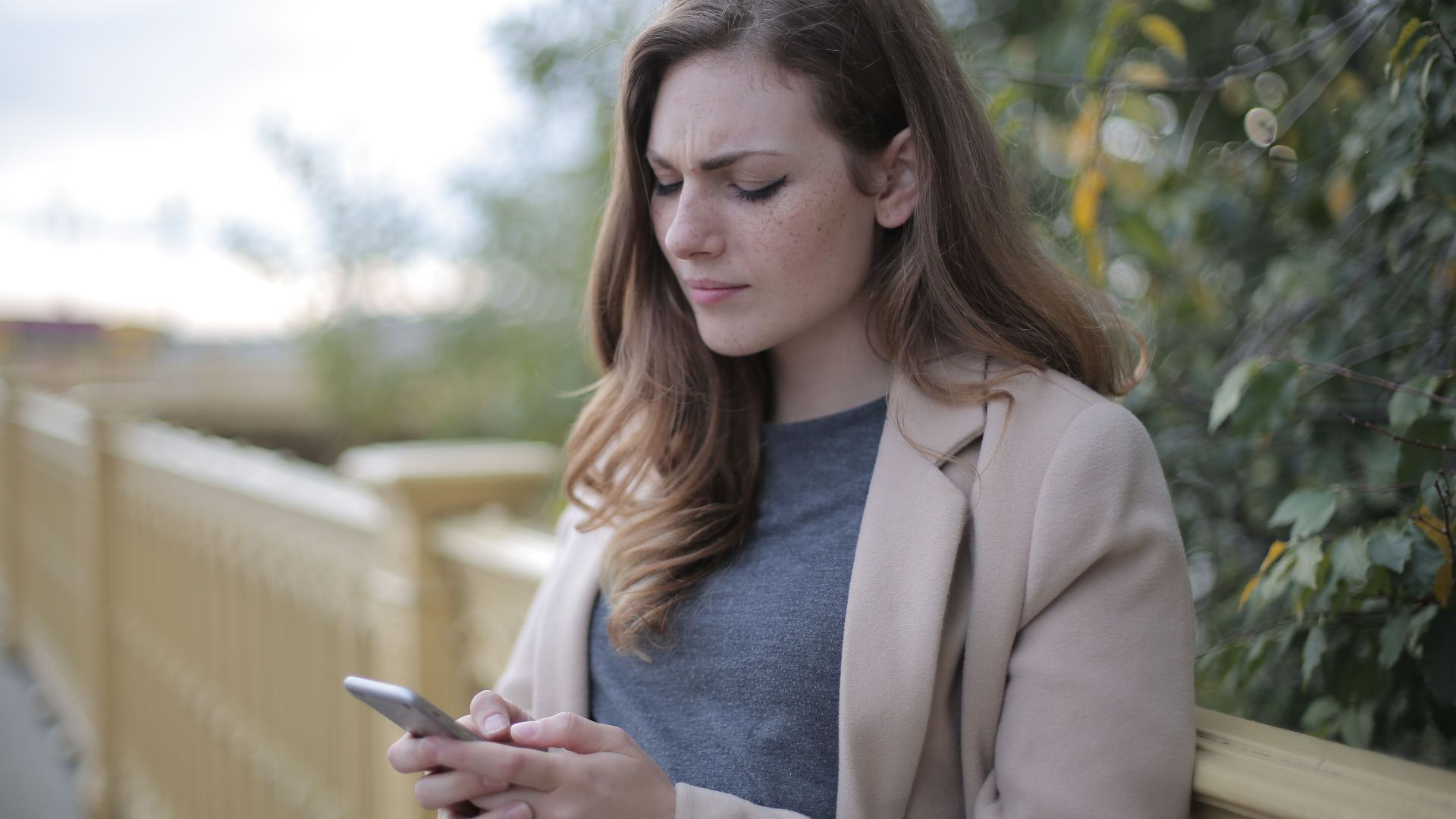 A woman stands outdoors in a casual outfit, using her smartphone with a pensive look.