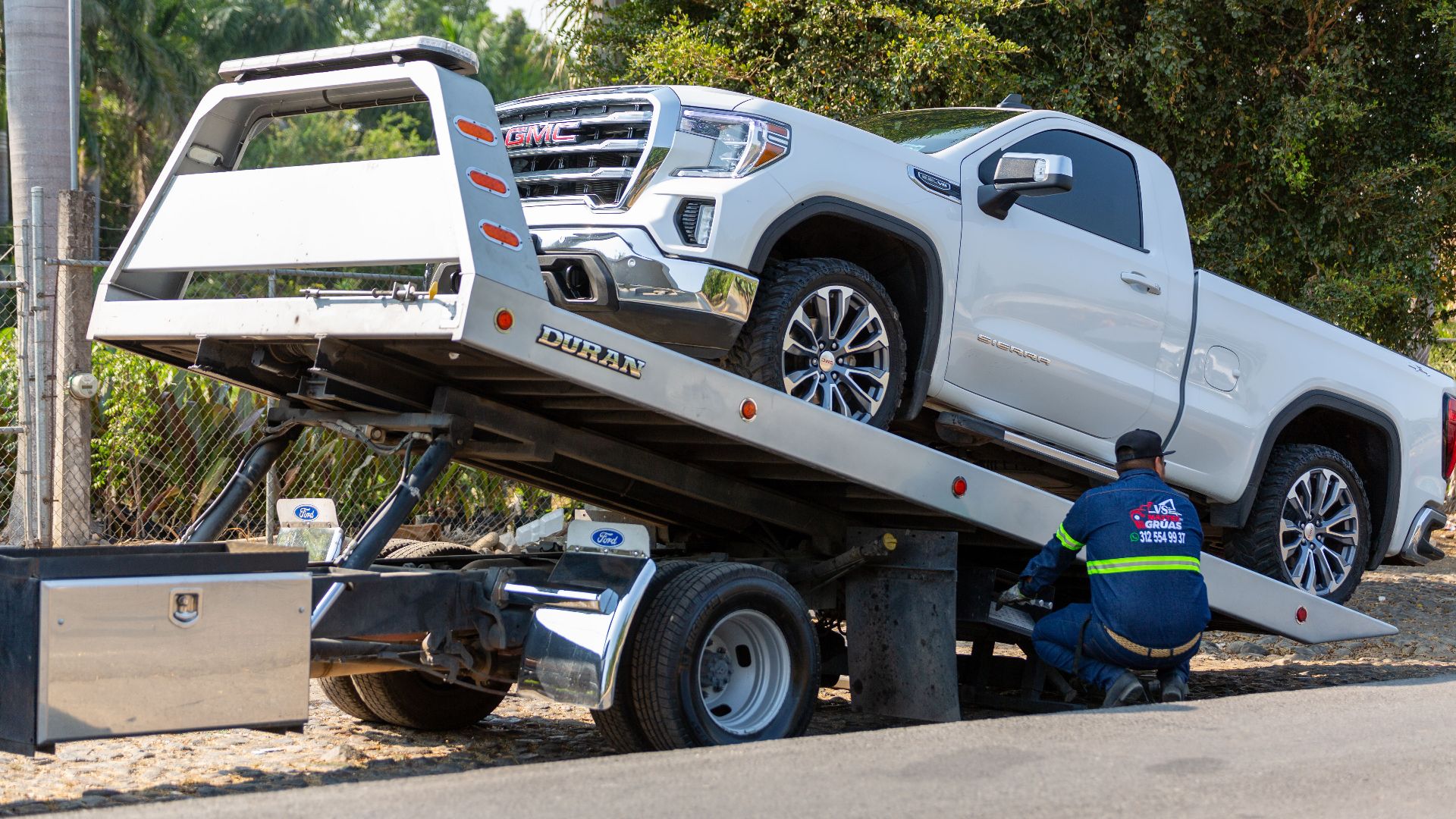 Tow truck operator loading white GMC pickup truck on street in daytime.