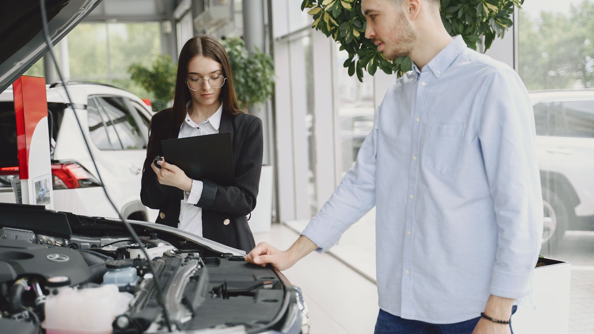 Man and woman examining car engine in dealership service area, focused on vehicle maintenance and functionality.
