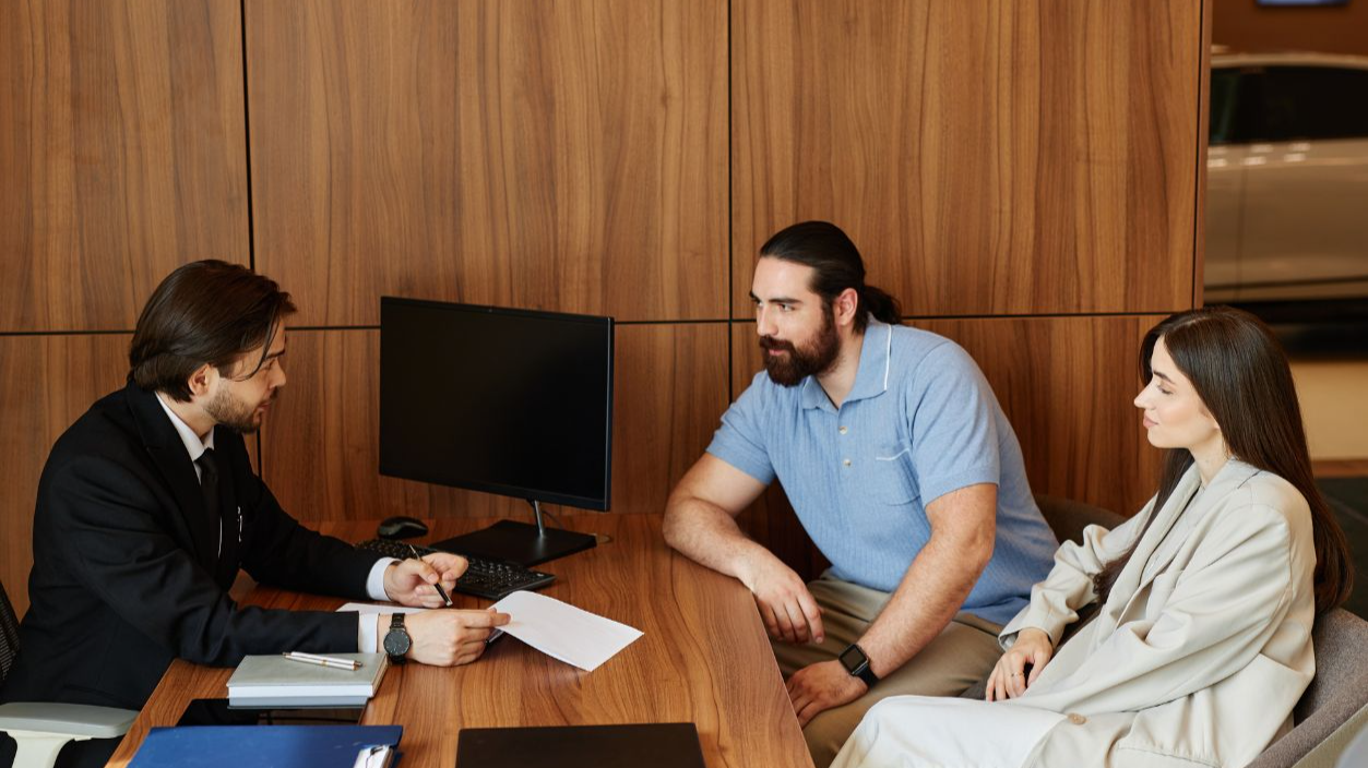 Shutterstock-2513248373, Side view of male car broker at desk making deal with couple buying new vehicle, discussing terms and conditions of contract at office of dealership center