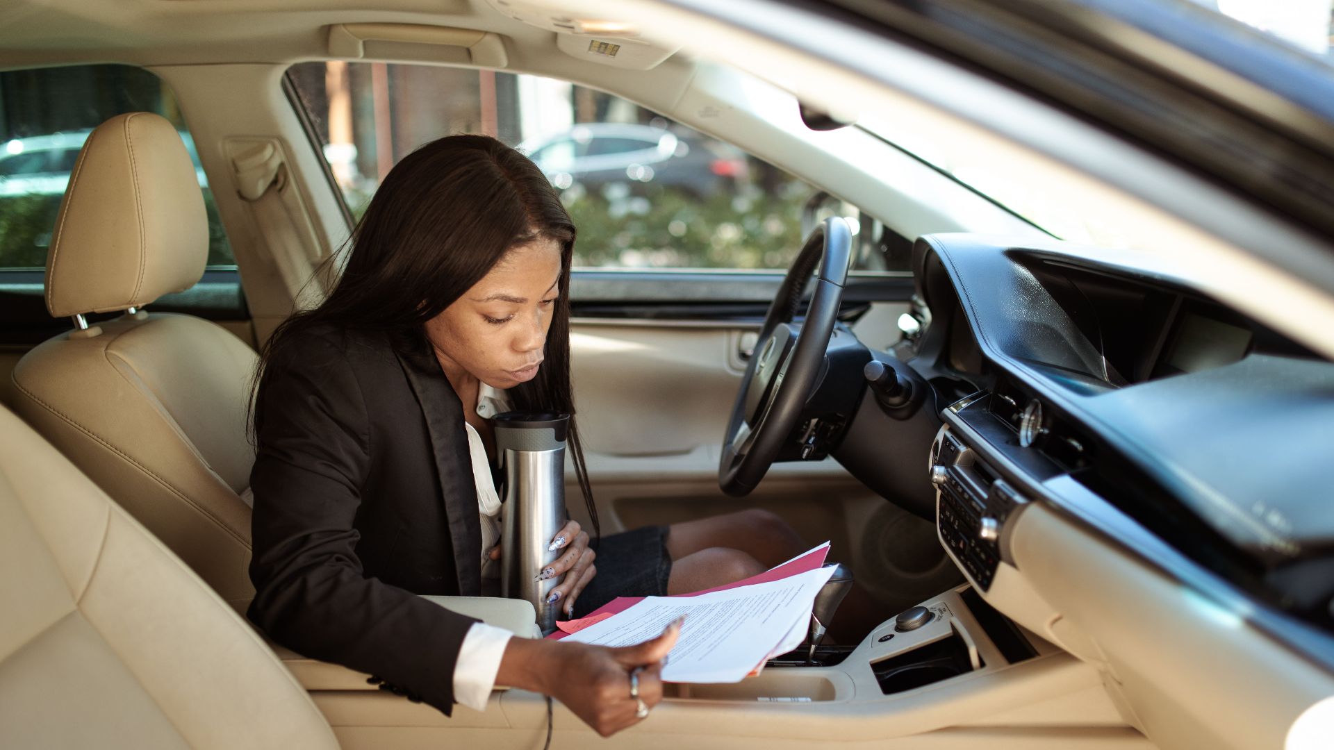 A focused businesswoman reads documents in her car, multitasking with a tumbler.