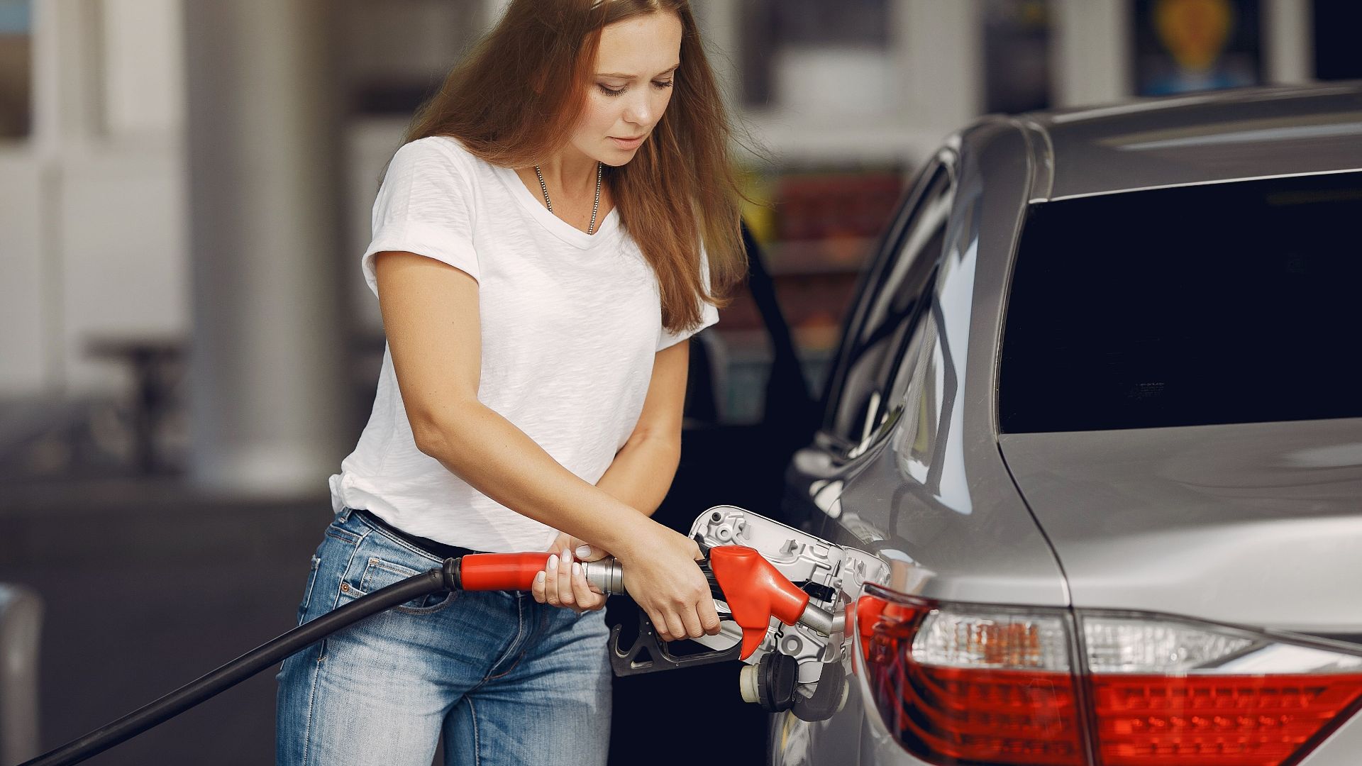 Attentive female driver in casual outfit and headband filling up modern automobile with automotive fuel gun on petrol station while looking down