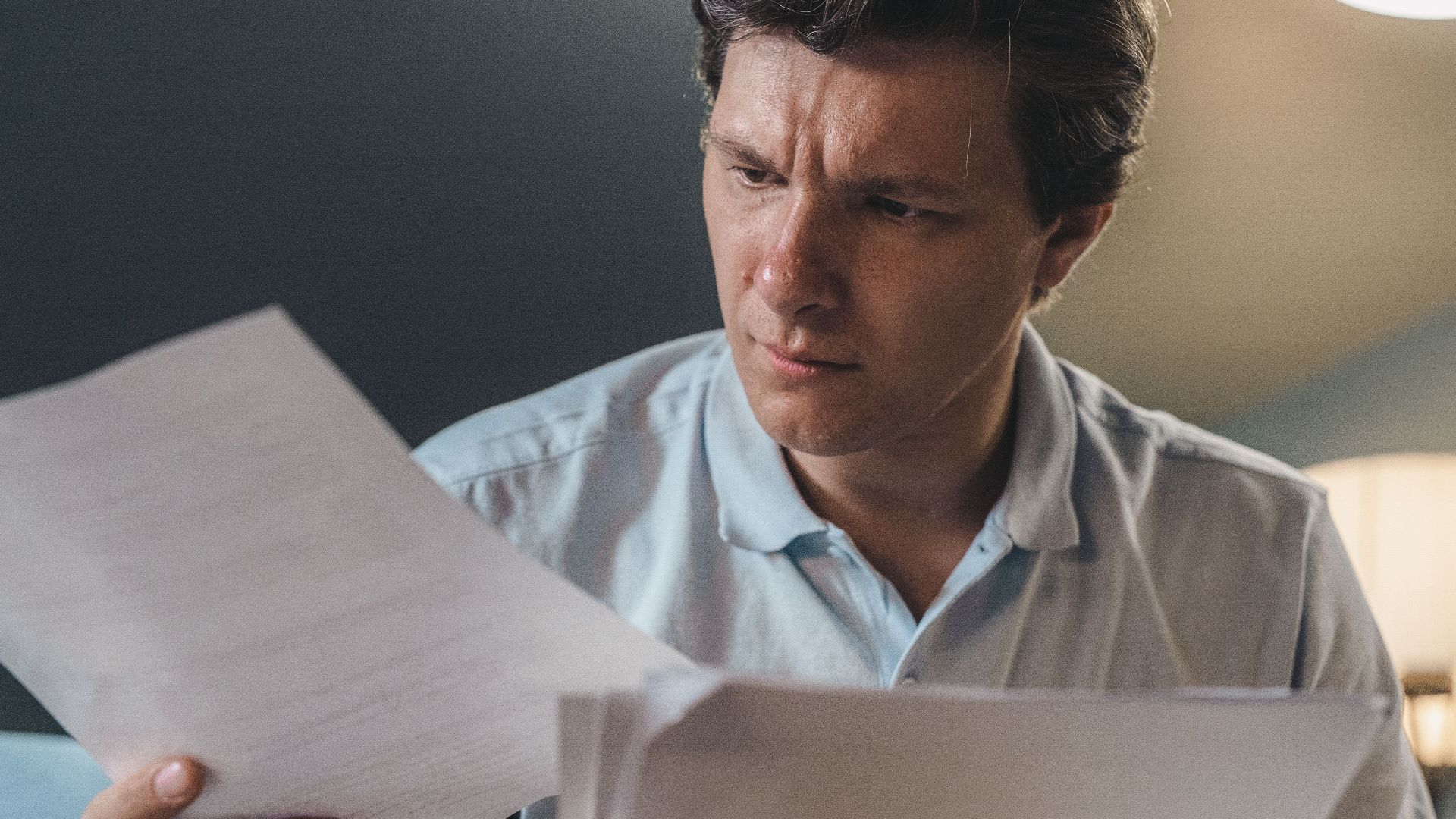 A man intently examines papers, seated indoors under warm lighting, focusing on his work.