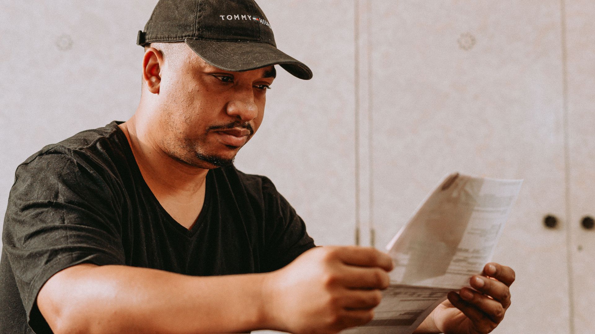 A man in a black cap reading papers at a table indoors.