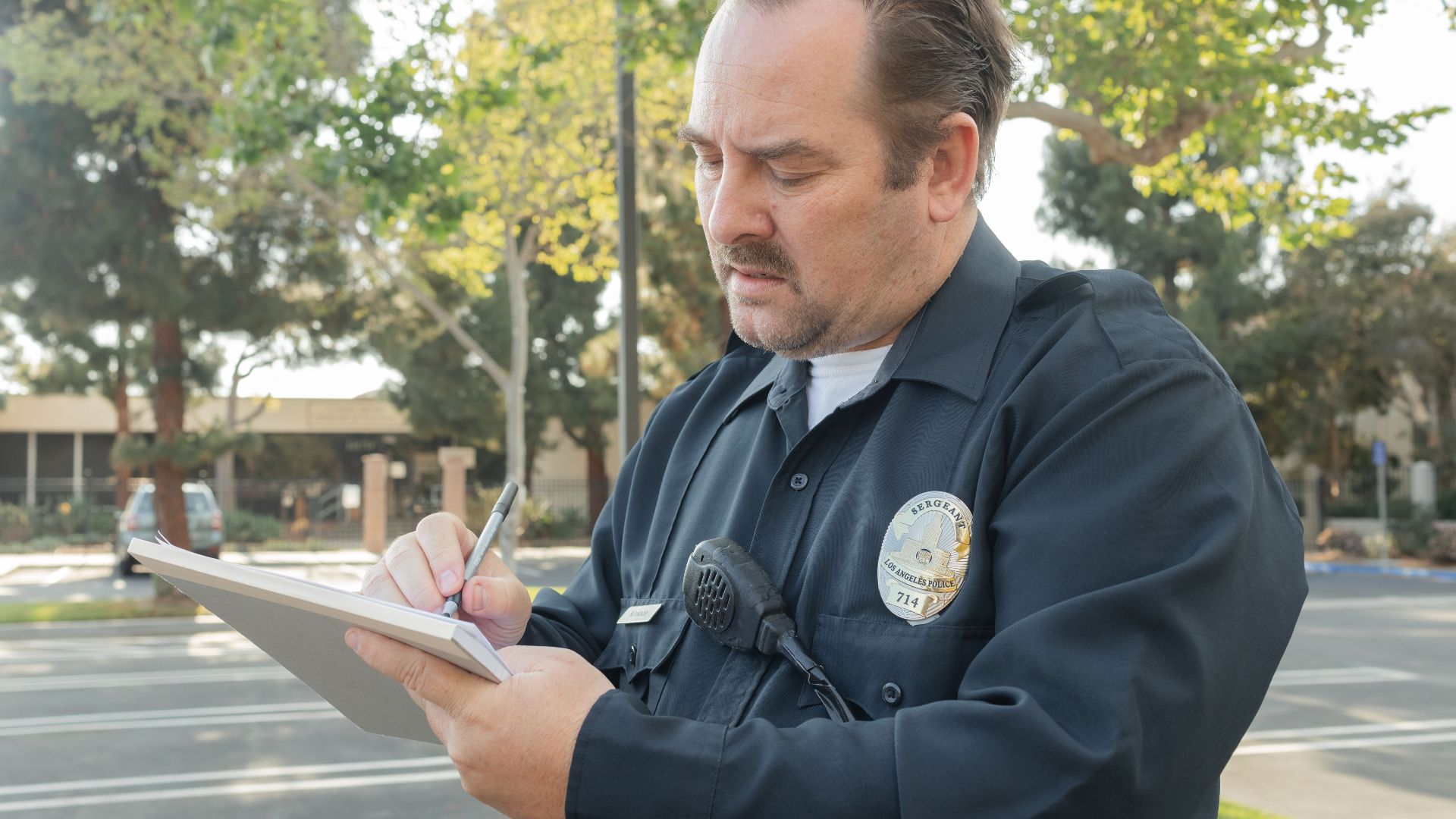 A police officer outdoors writing a report on a notepad. Trees in the background.