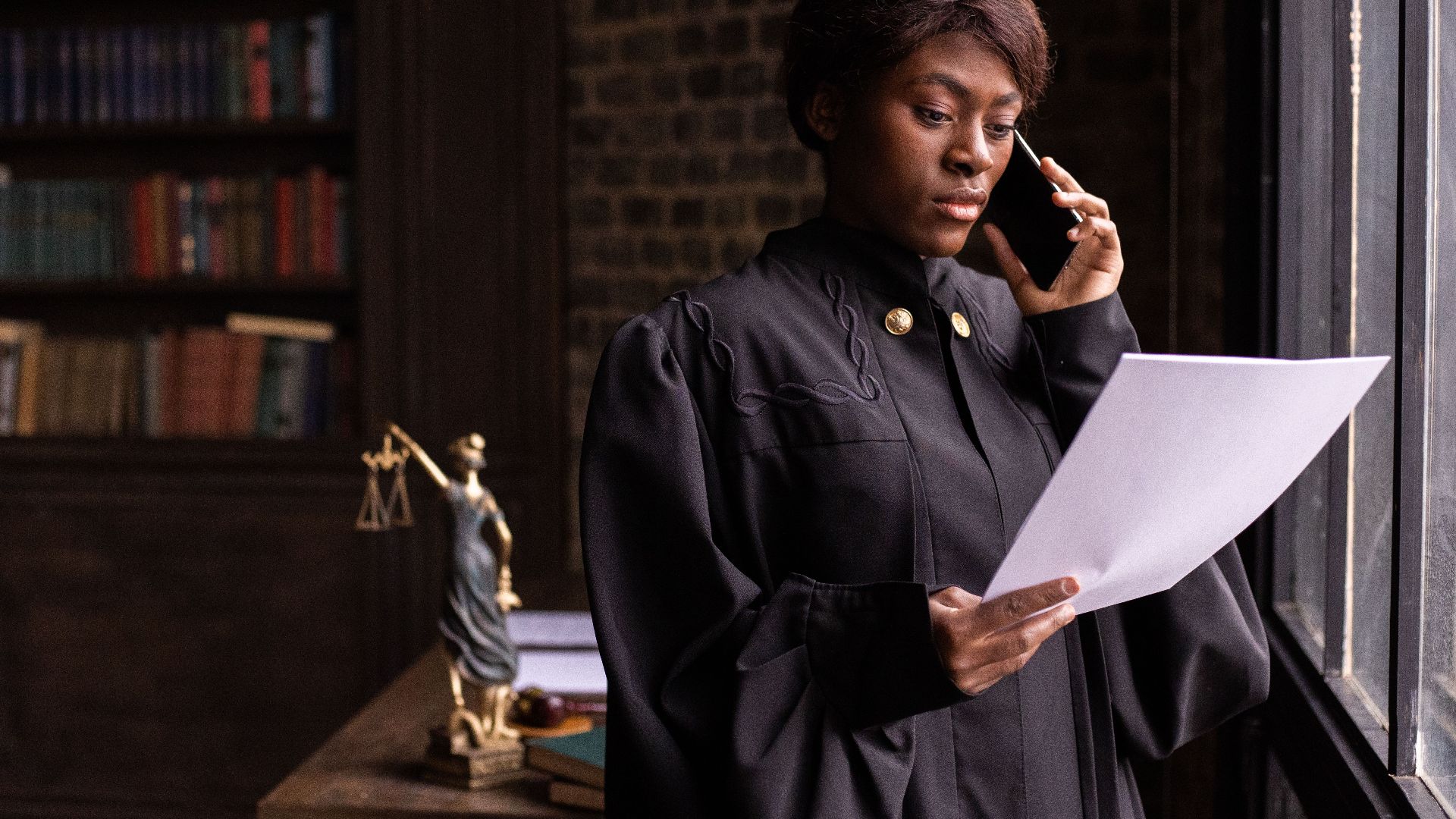 African American judge in robe, reading document and talking on phone by the office window.