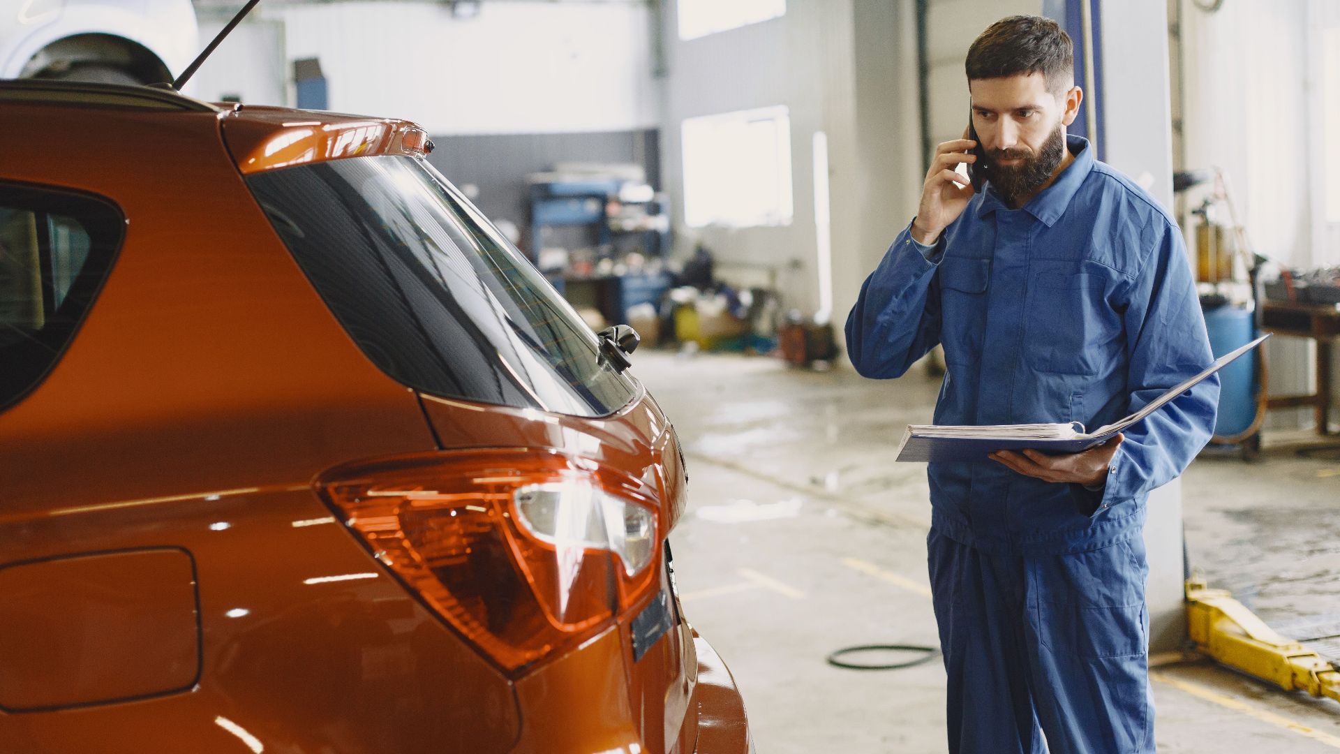 Mechanic in a blue uniform talks on phone beside a red car in an auto repair shop.