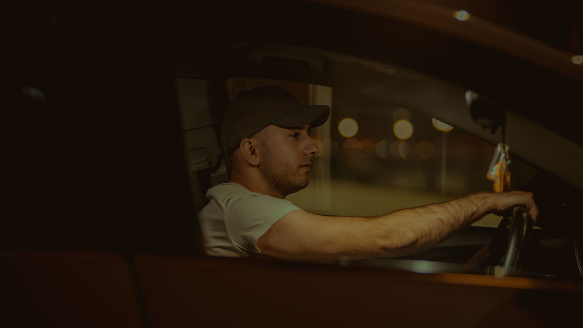 A man wearing a cap is driving a car at night, focused on the road ahead.