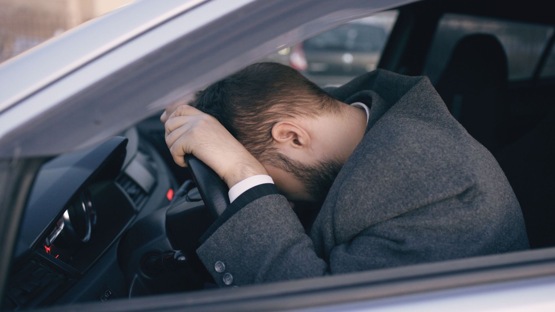 Man feeling overwhelmed and stressed, resting his head on a steering wheel inside a car.