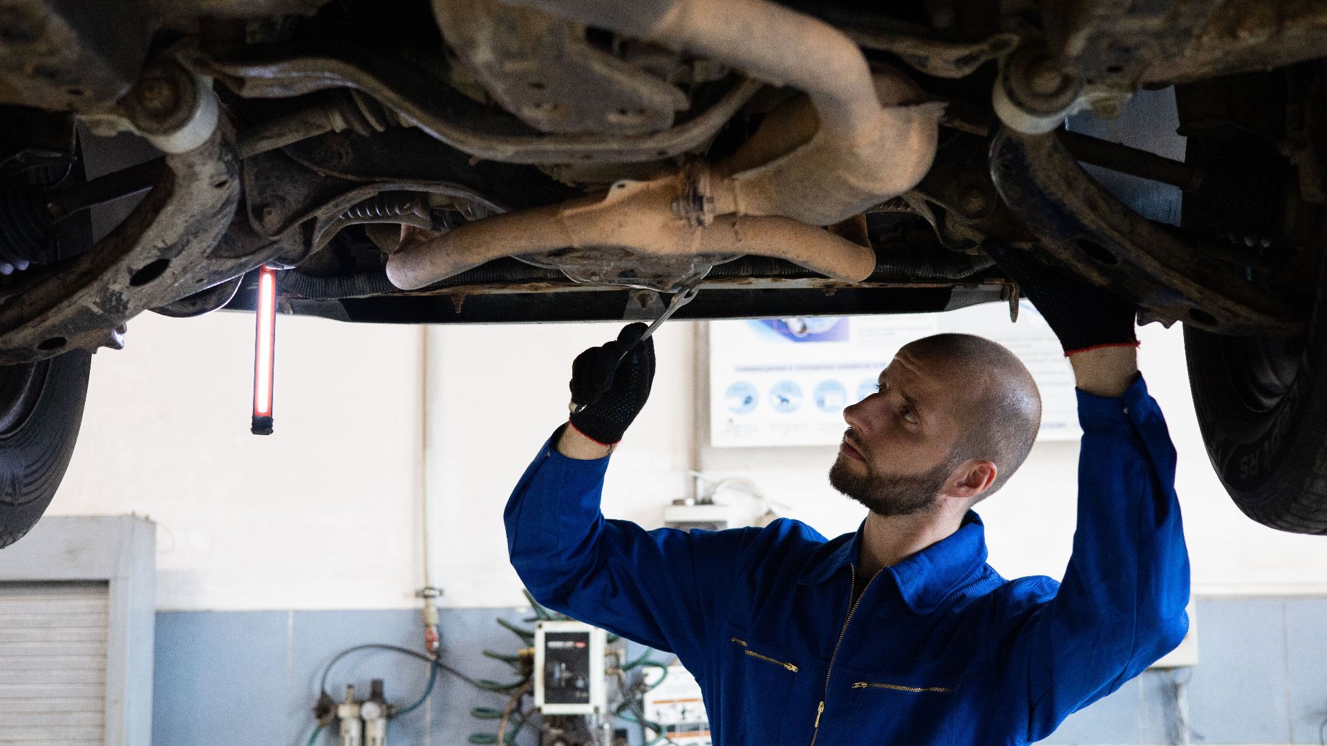 A mechanic in blue overalls inspects the exhaust system of a car in a workshop.