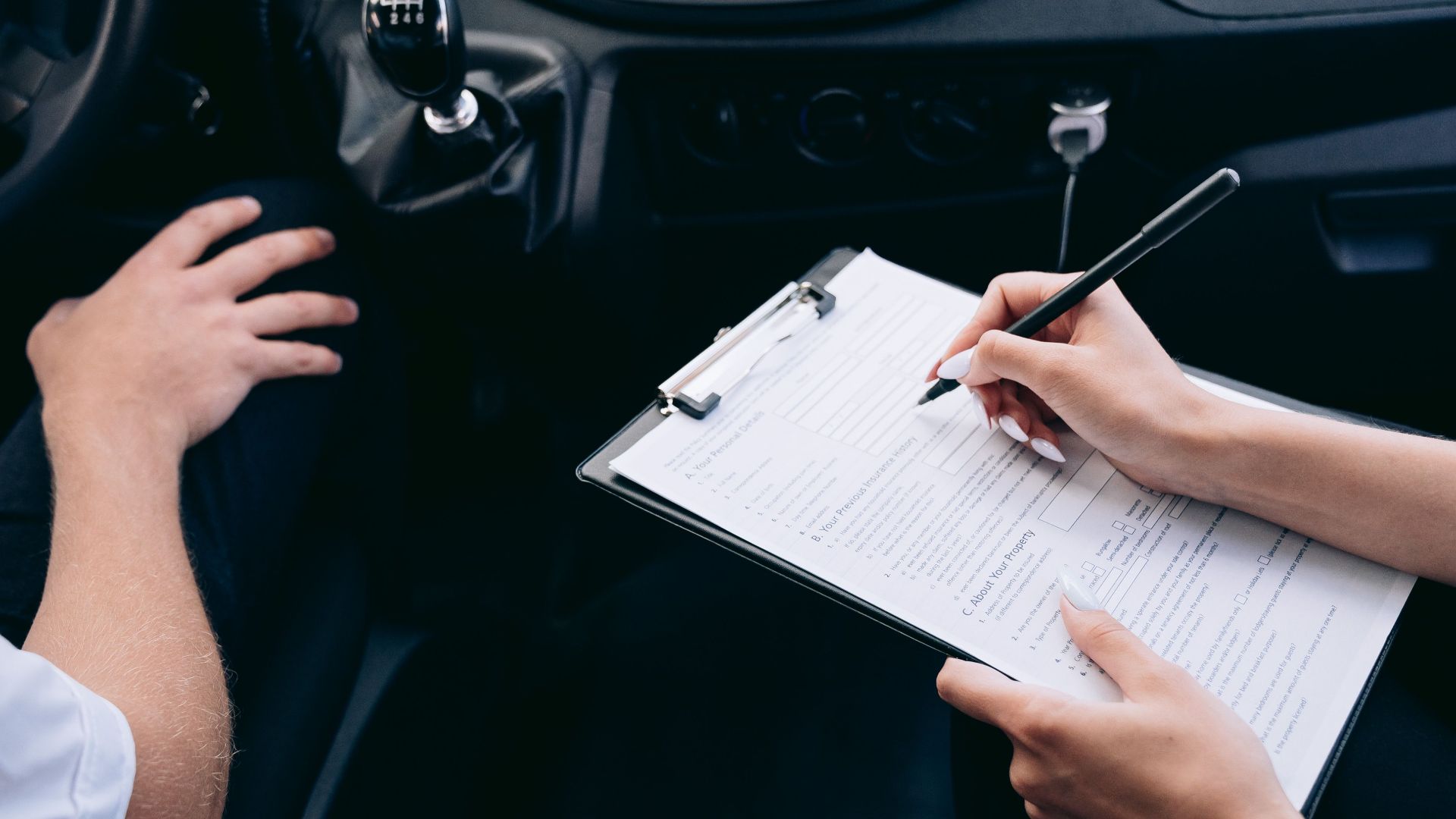 Close-up of a person writing on a clipboard inside a car, showing hands and a gear shift.