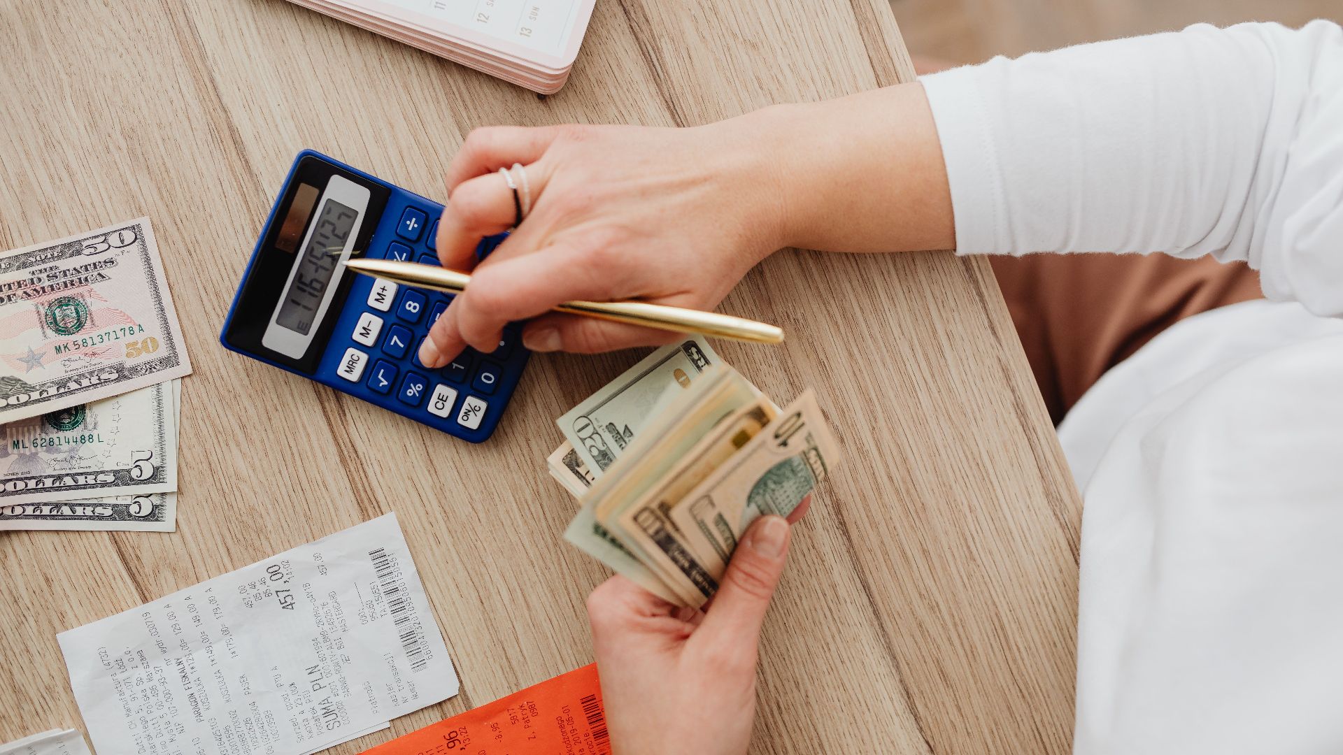 Person using a calculator and counting cash on a wooden table with receipts.