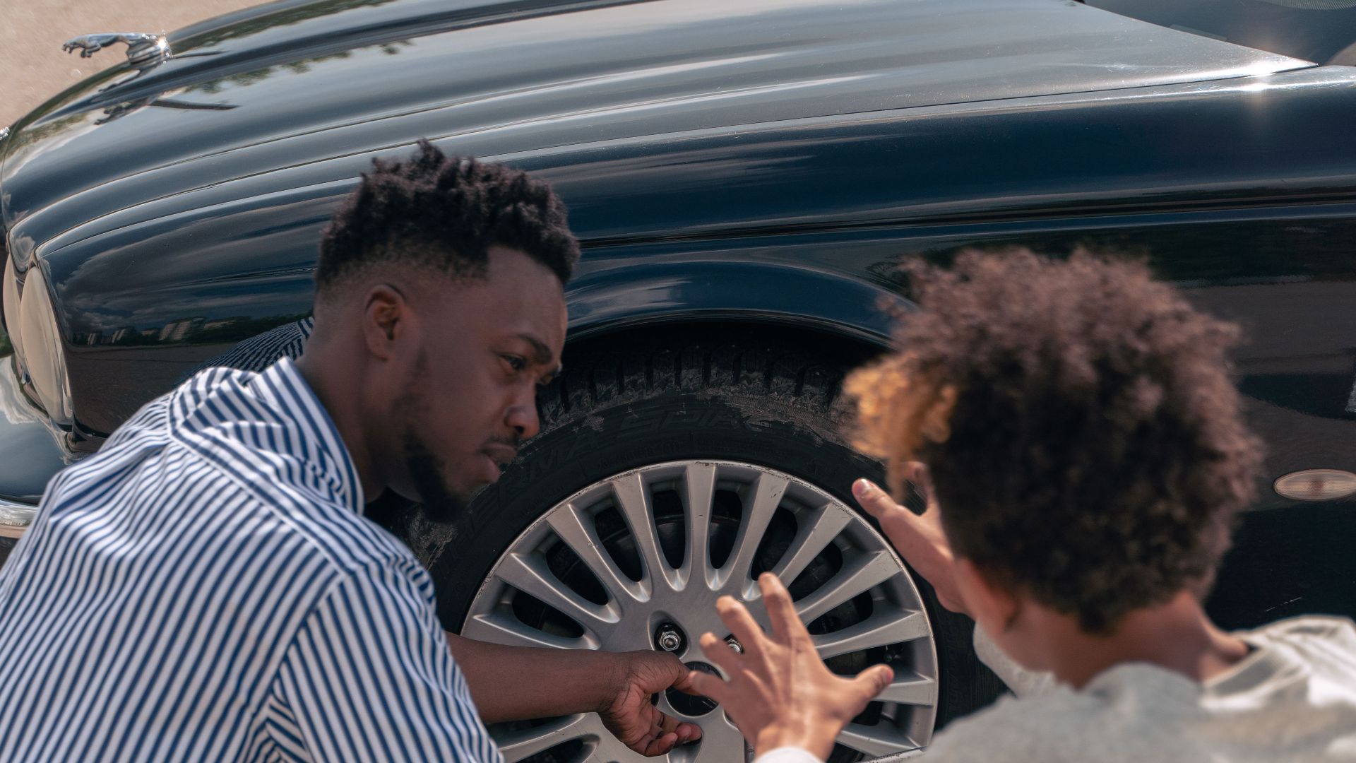 A father instructs his teenage son on how to change a car tire outdoors.