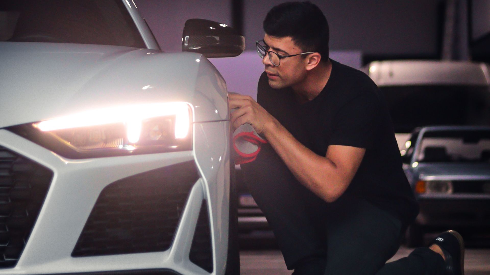Man kneeling beside a white sports car, inspecting the headlight in a garage.