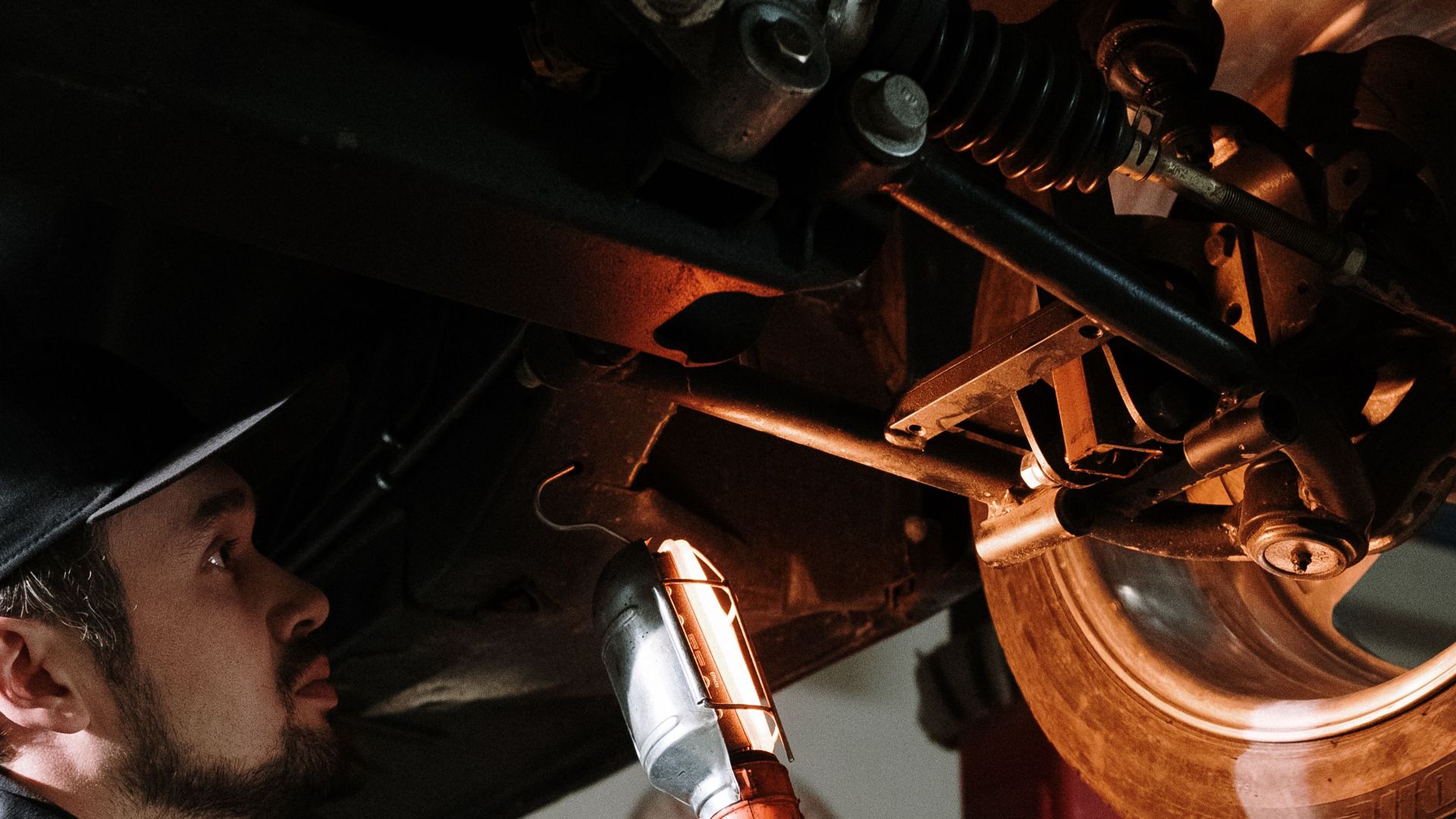Mechanic uses a flashlight to inspect car's suspension in a garage setting.