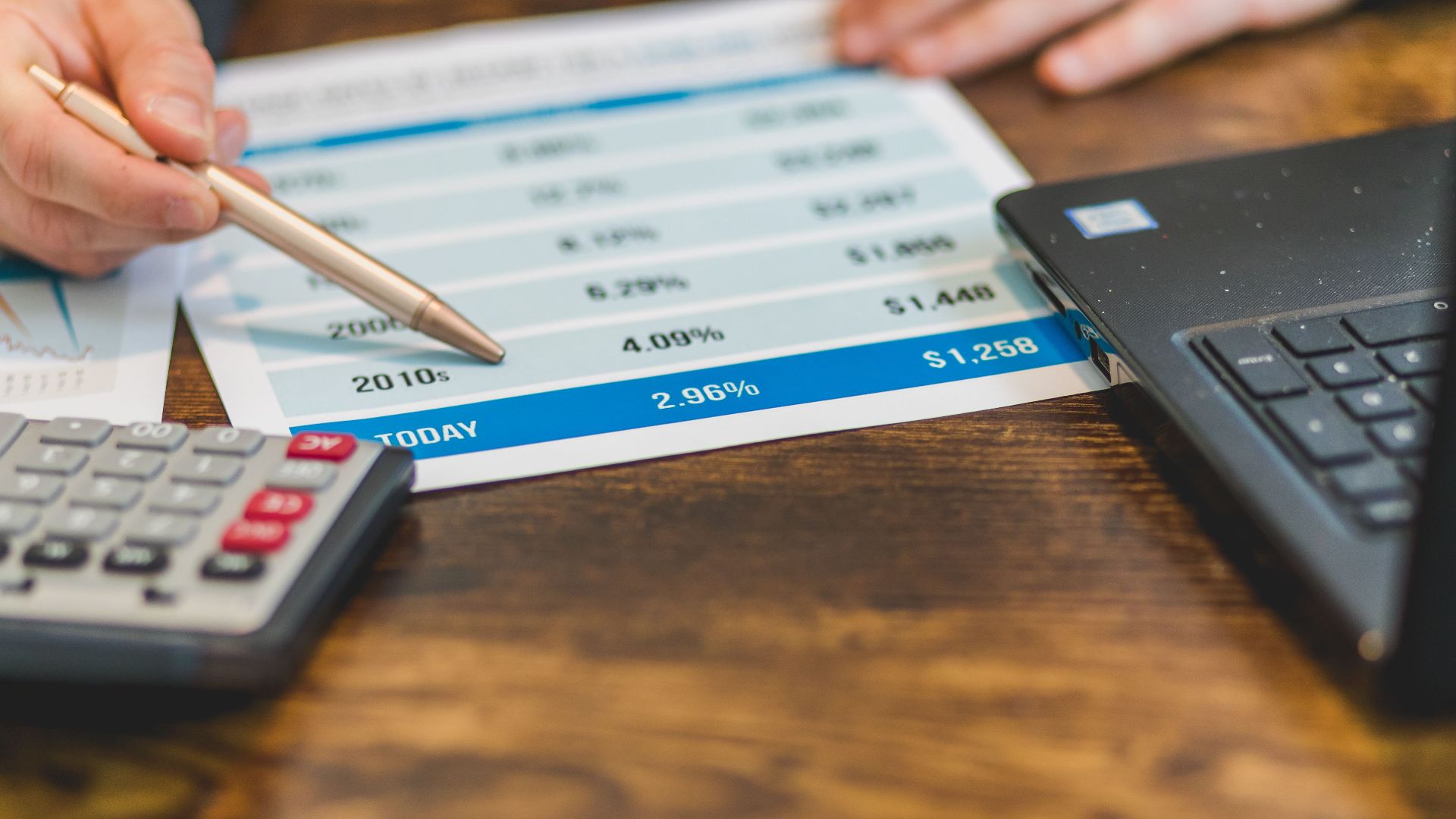 Close-up of a hand with pen analyzing financial rates on paper with a calculator and laptop nearby.