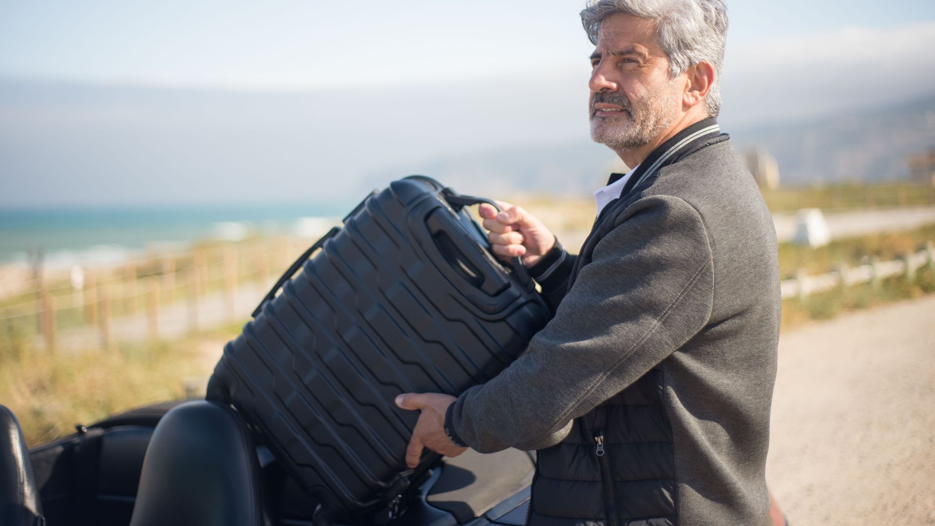 Elderly man with suitcase by a convertible on a scenic coastal journey in Portugal.