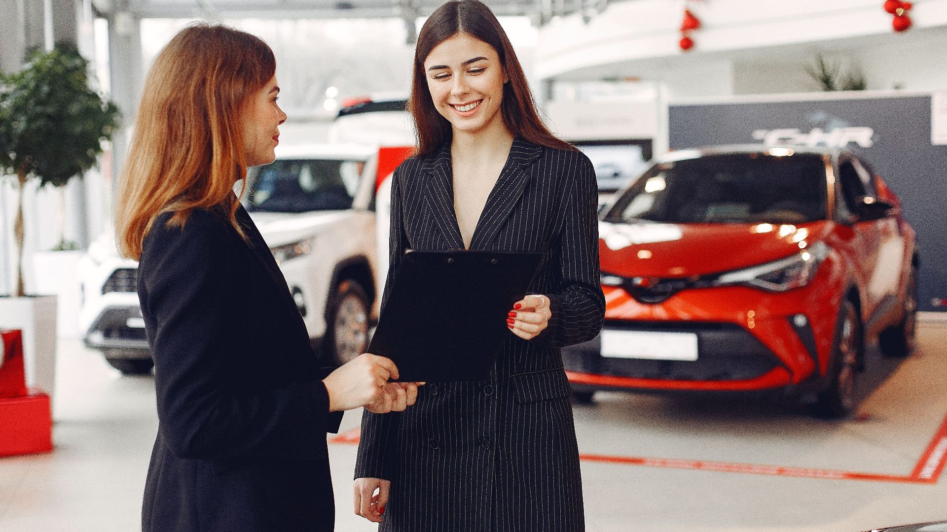 Cheerful young friendly dealer in formal stylish black dress showing contract to smiling female customer in black jacket while standing in car showroom against new shiny automobiles