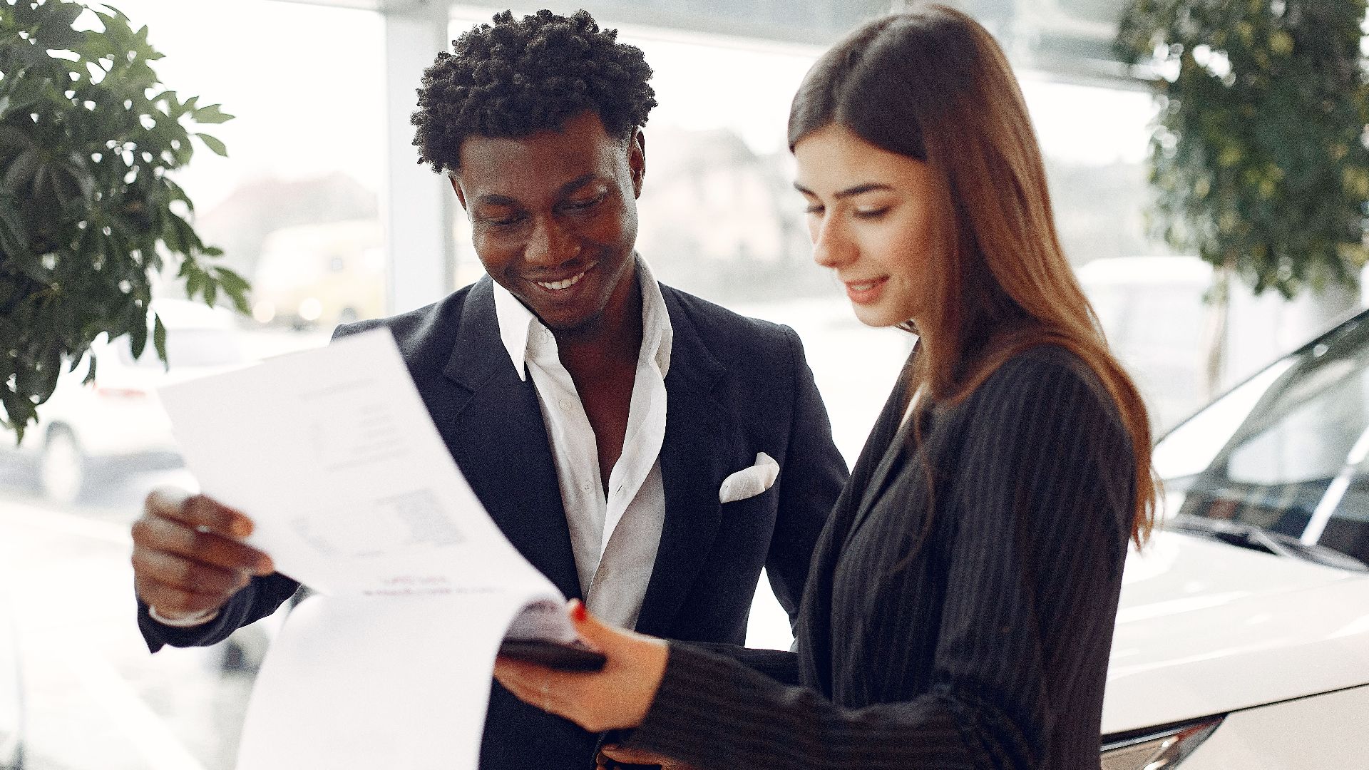 Cheerful smiling multiethnic businessman in classy suit and professional elegant female consultant standing close and reading contract details in car showroom