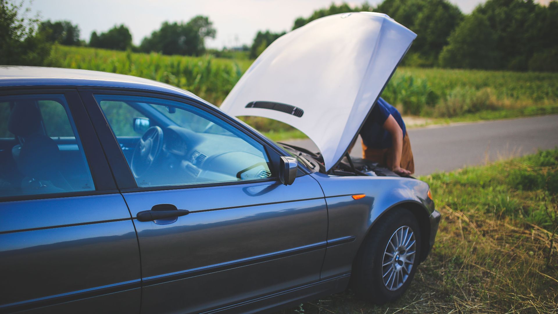 Man checking car engine with hood open by the side of a rural road.