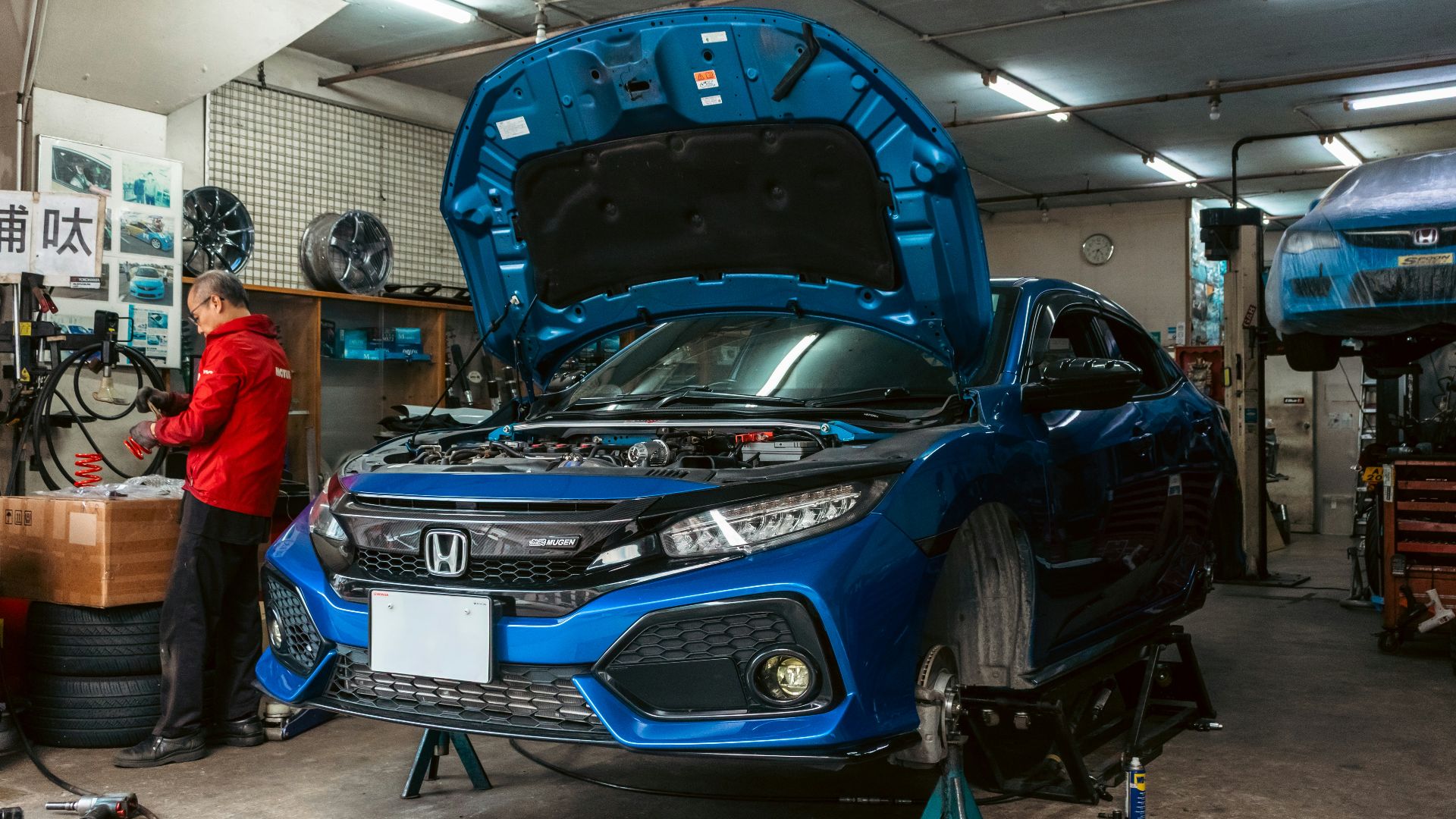 A mechanic in a red jacket working on a blue car in a garage