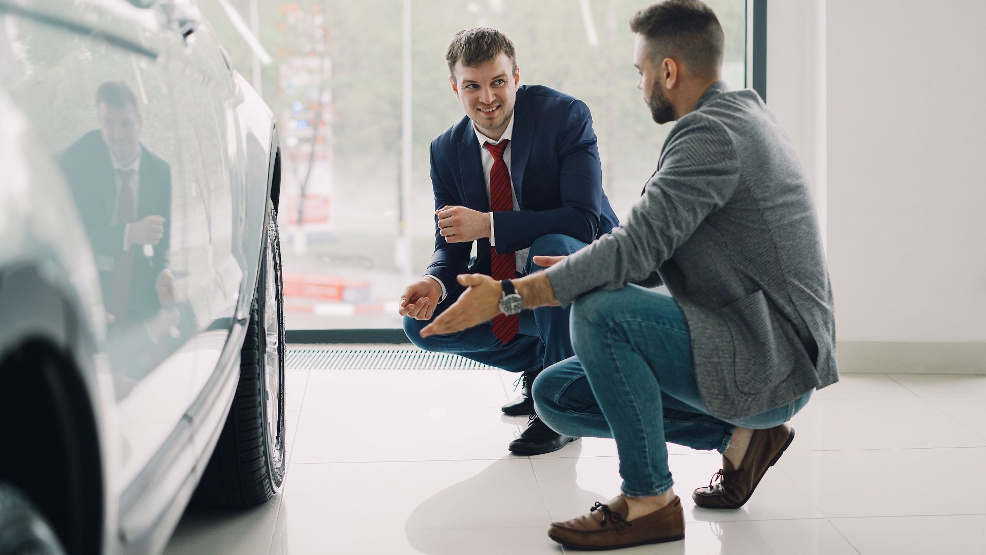 Two men discussing car features in a showroom, kneeling near a vehicle.