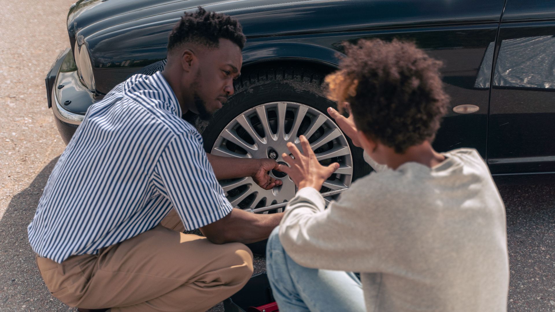 A father instructs his teenage son on how to change a car tire outdoors.